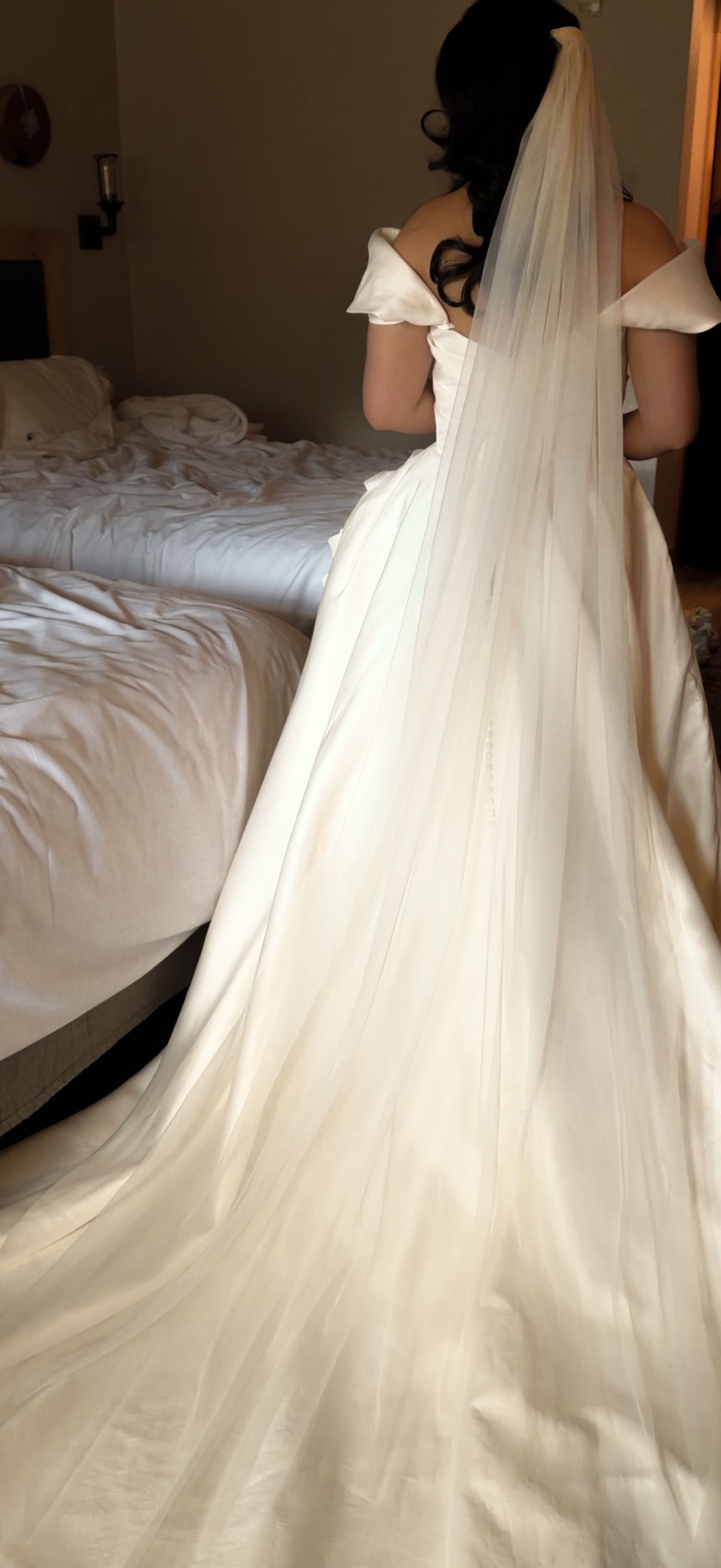 Bride in off-shoulder gown with long cathedral veil trailing behind her in a hotel room before wedding.