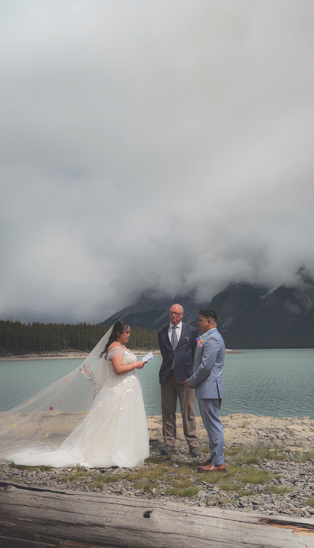 Couple exchanges vows lakeside with officiant under cloudy mountain sky at intimate outdoor ceremony.