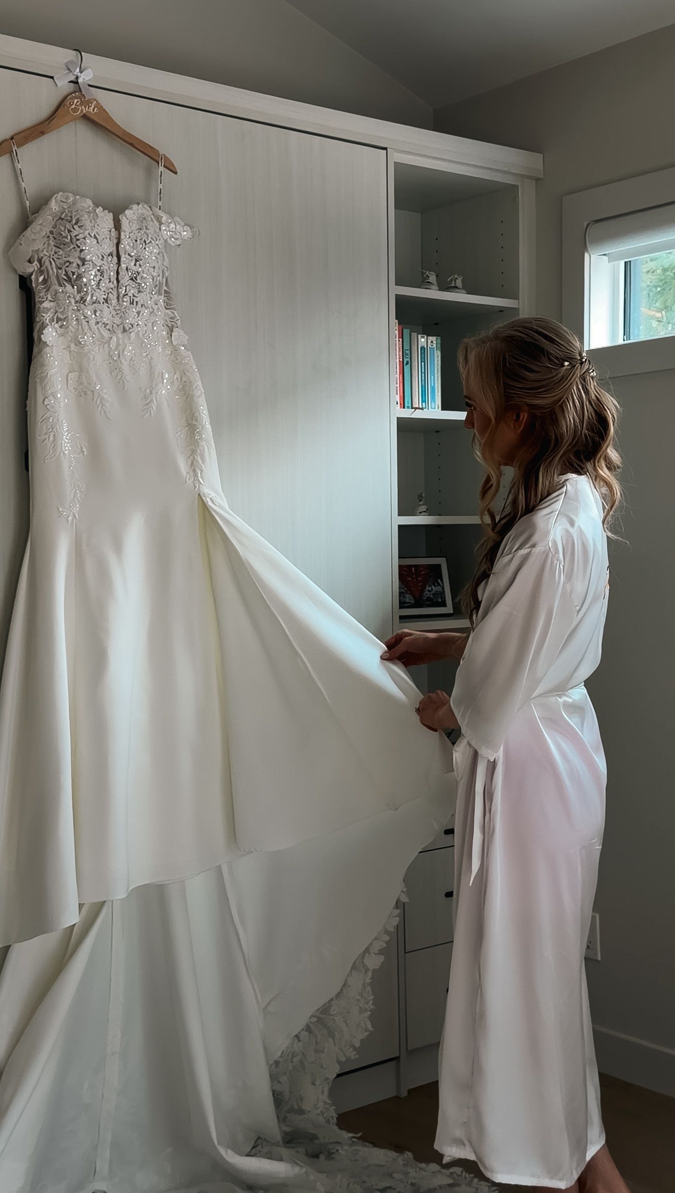Bride in white robe admires her lace and satin wedding gown hanging in a bright room.