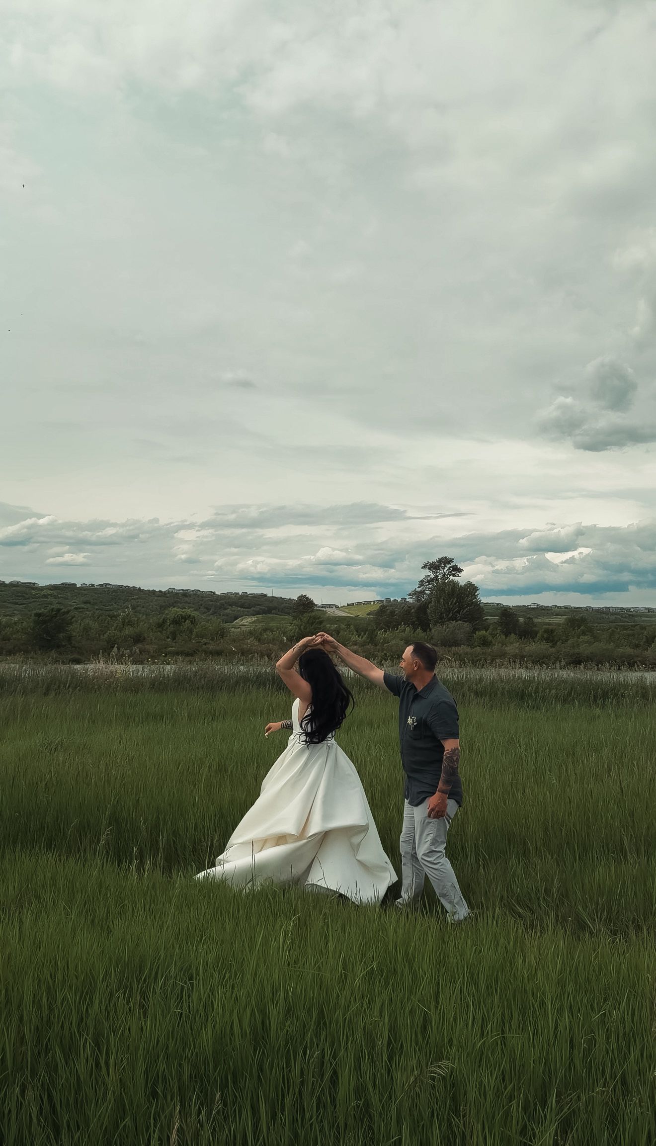 A groom spins his bride in a white wedding gown through a vibrant green meadow under cloudy skies.