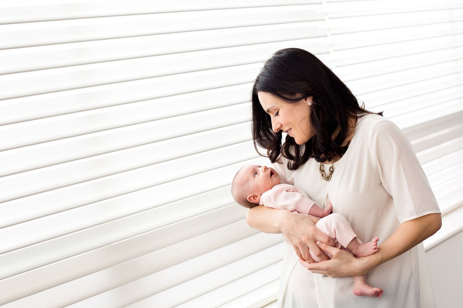 A dark-haired mother in a white dress tenderly gazes at her newborn baby by a bright window.