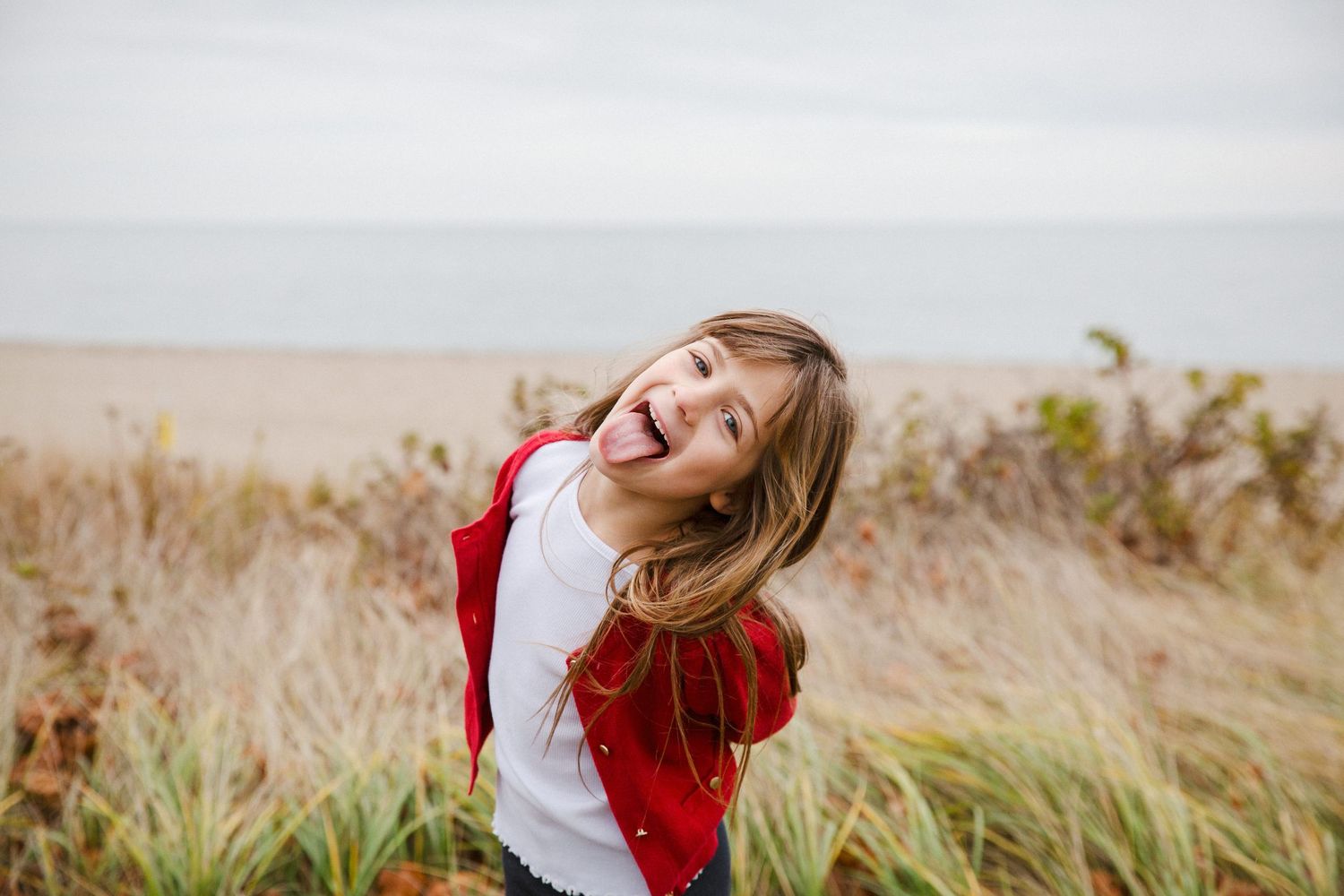 Playful girl in red jacket sticking out tongue while standing in coastal grass on overcast day.