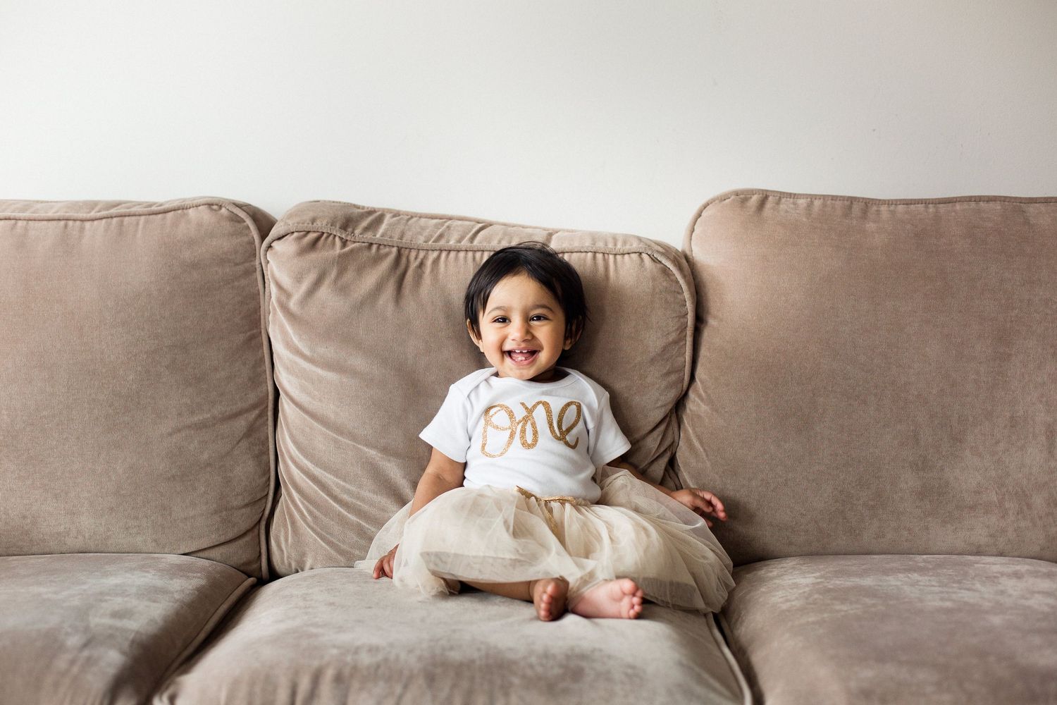 Happy toddler laughing while sitting on a beige sofa wearing a white one birthday shirt.
