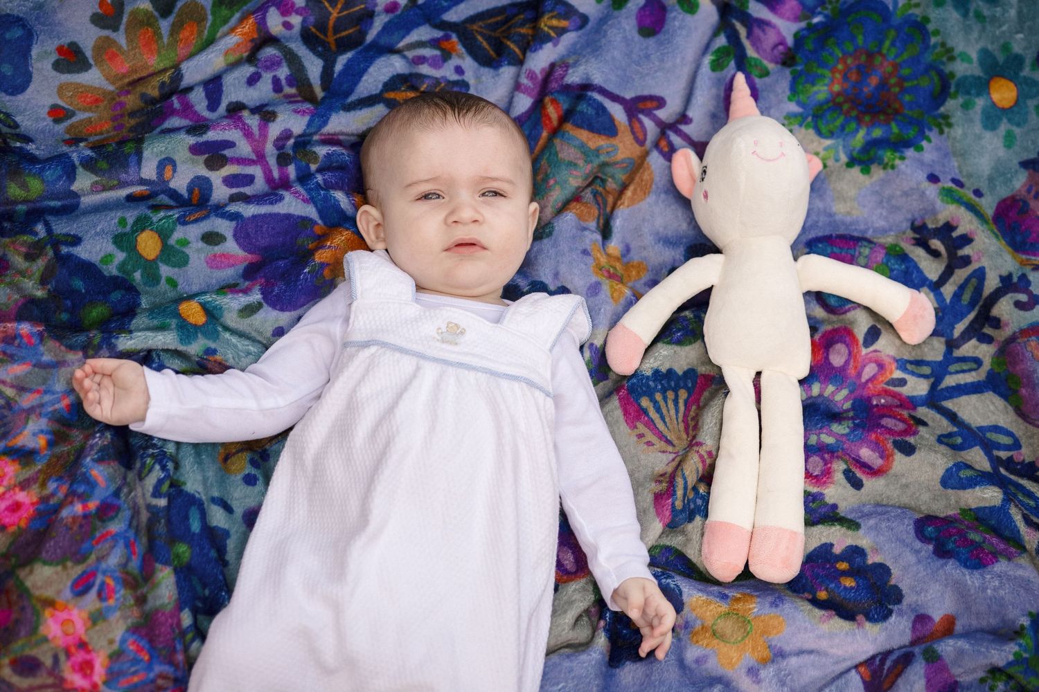 Adorable baby in white outfit lying on vibrant floral blanket next to a soft unicorn stuffed toy.