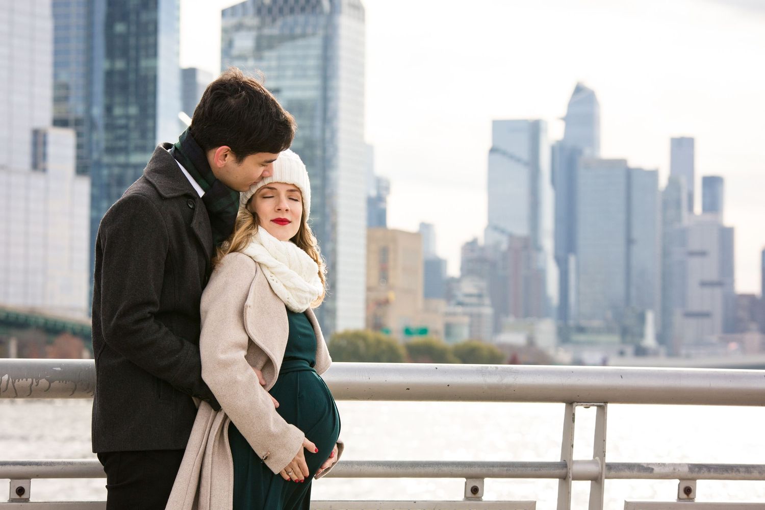 Pregnant couple embracing on waterfront with NYC skyline backdrop, wearing winter coats and white knit hat.