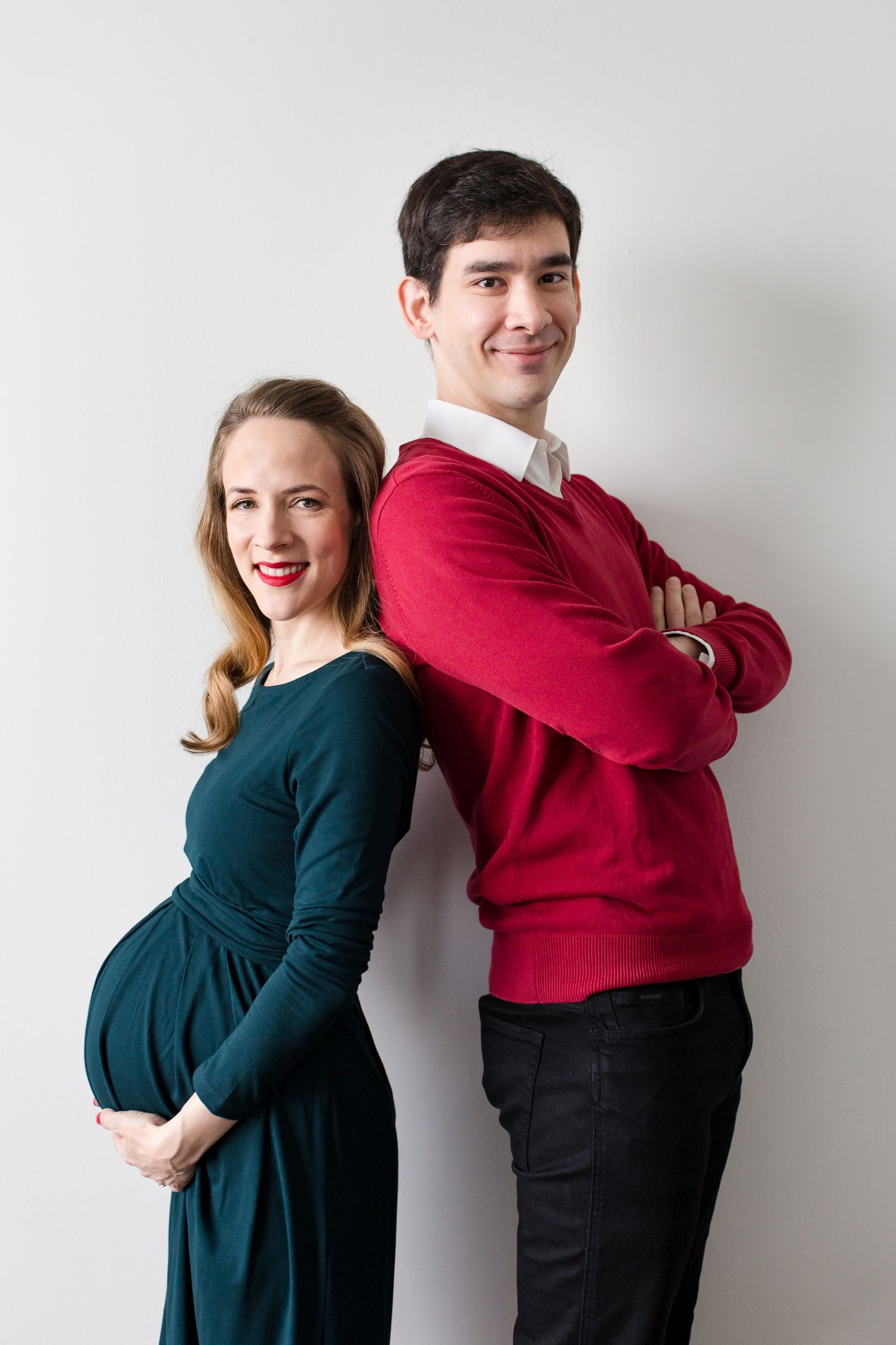 Smiling pregnant woman in teal dress and man in red sweater posing back-to-back against white background.