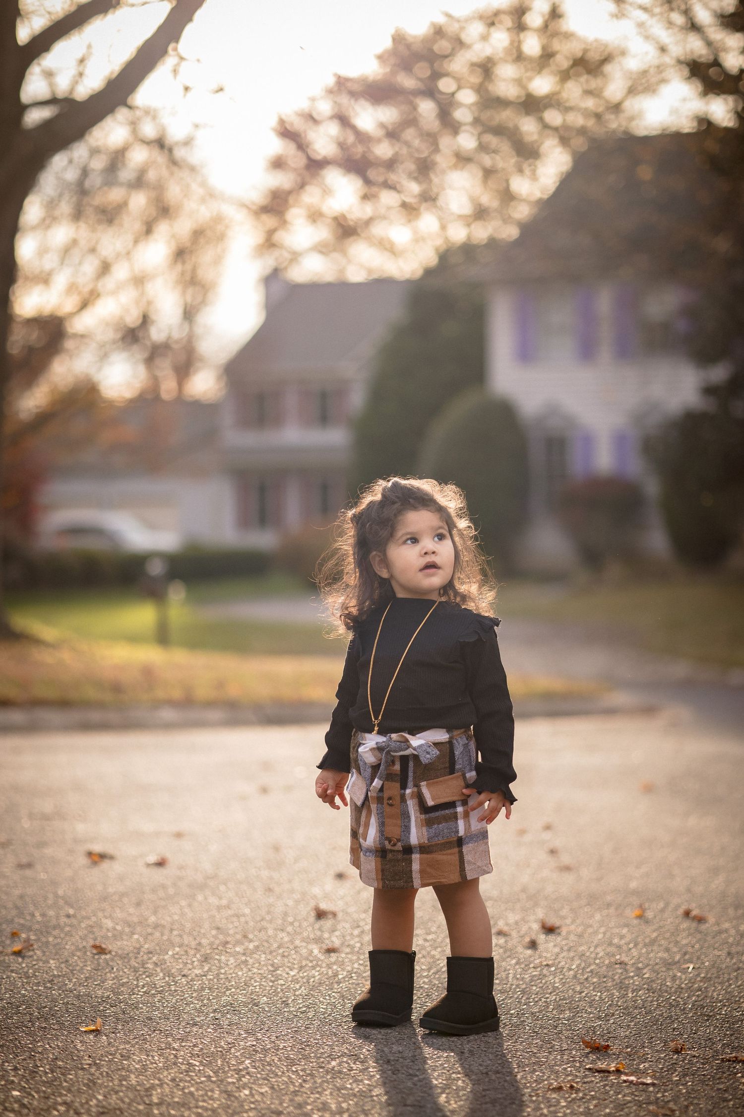 Toddler girl in plaid skirt and black turtleneck stands on suburban street during golden hour fall afternoon.