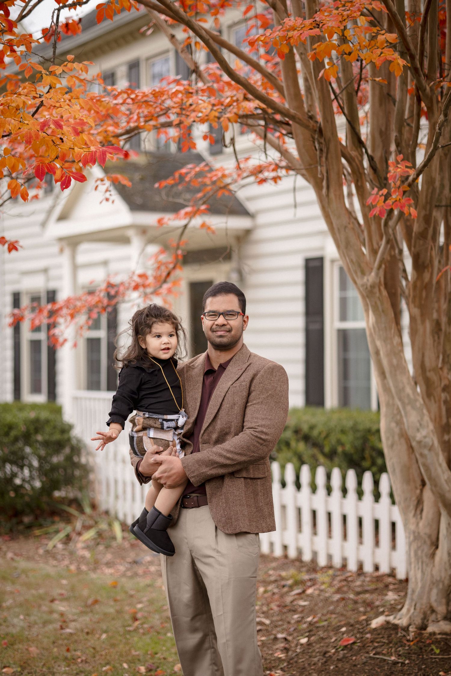 Father holding young child outdoors in autumn, standing near red-leafed tree beside white picket fence and house.