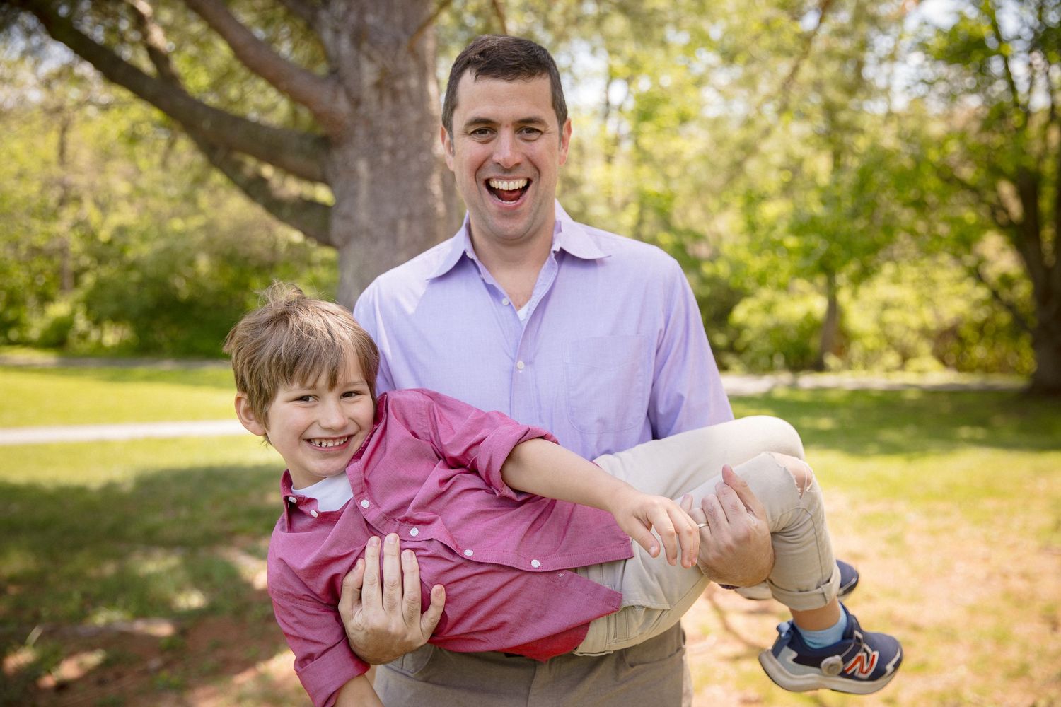 Man laughing while holding smiling boy in park with green trees in background on sunny day.