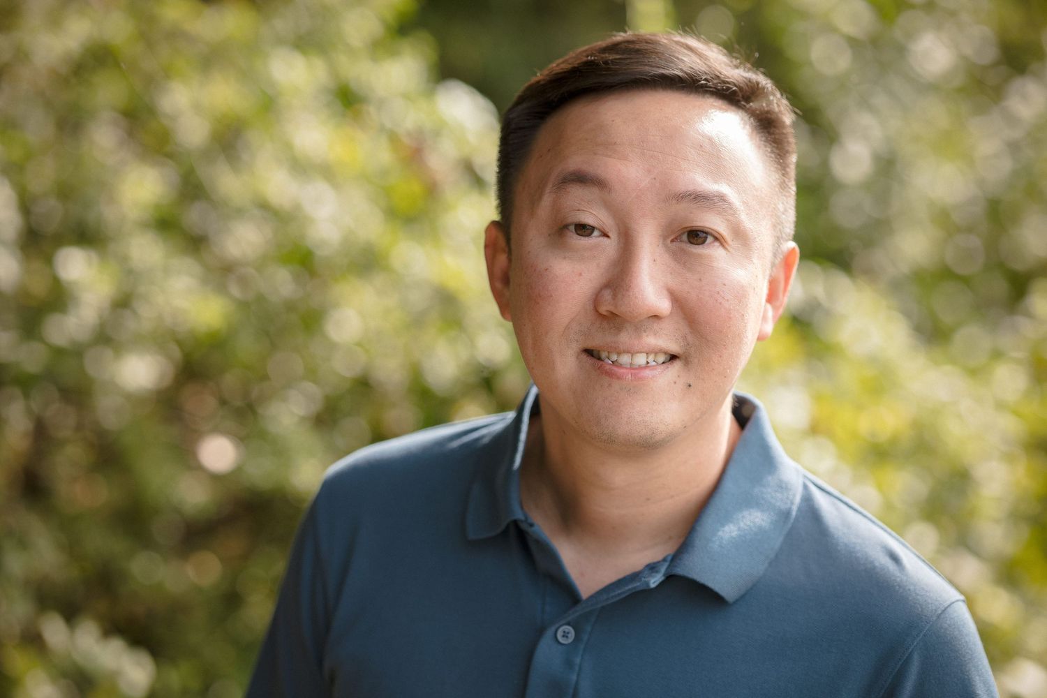 Smiling Asian man in blue polo shirt poses outdoors with green bokeh foliage background in professional headshot.