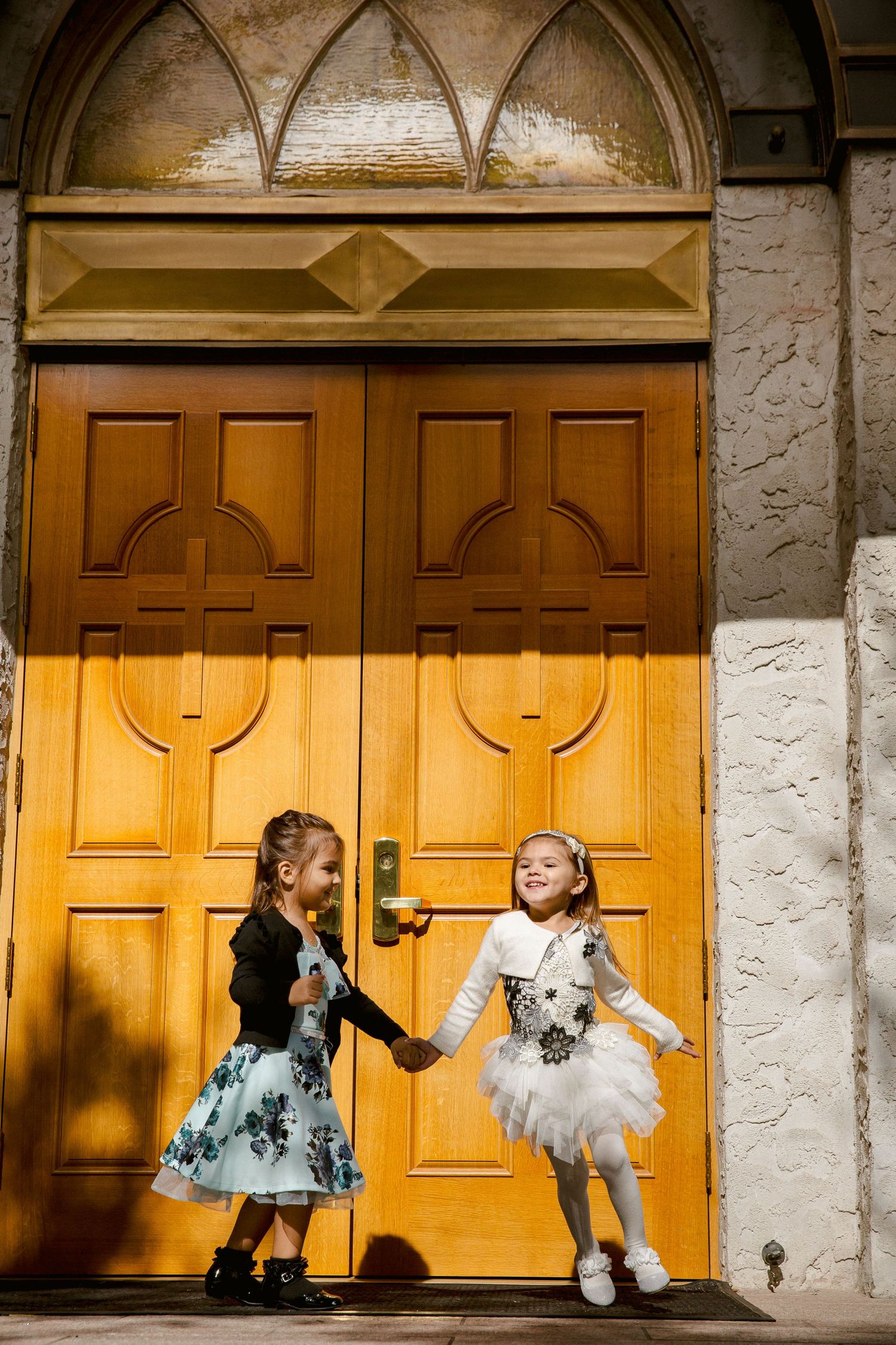 Two little girls in dresses holding hands and dancing in front of ornate wooden doors.