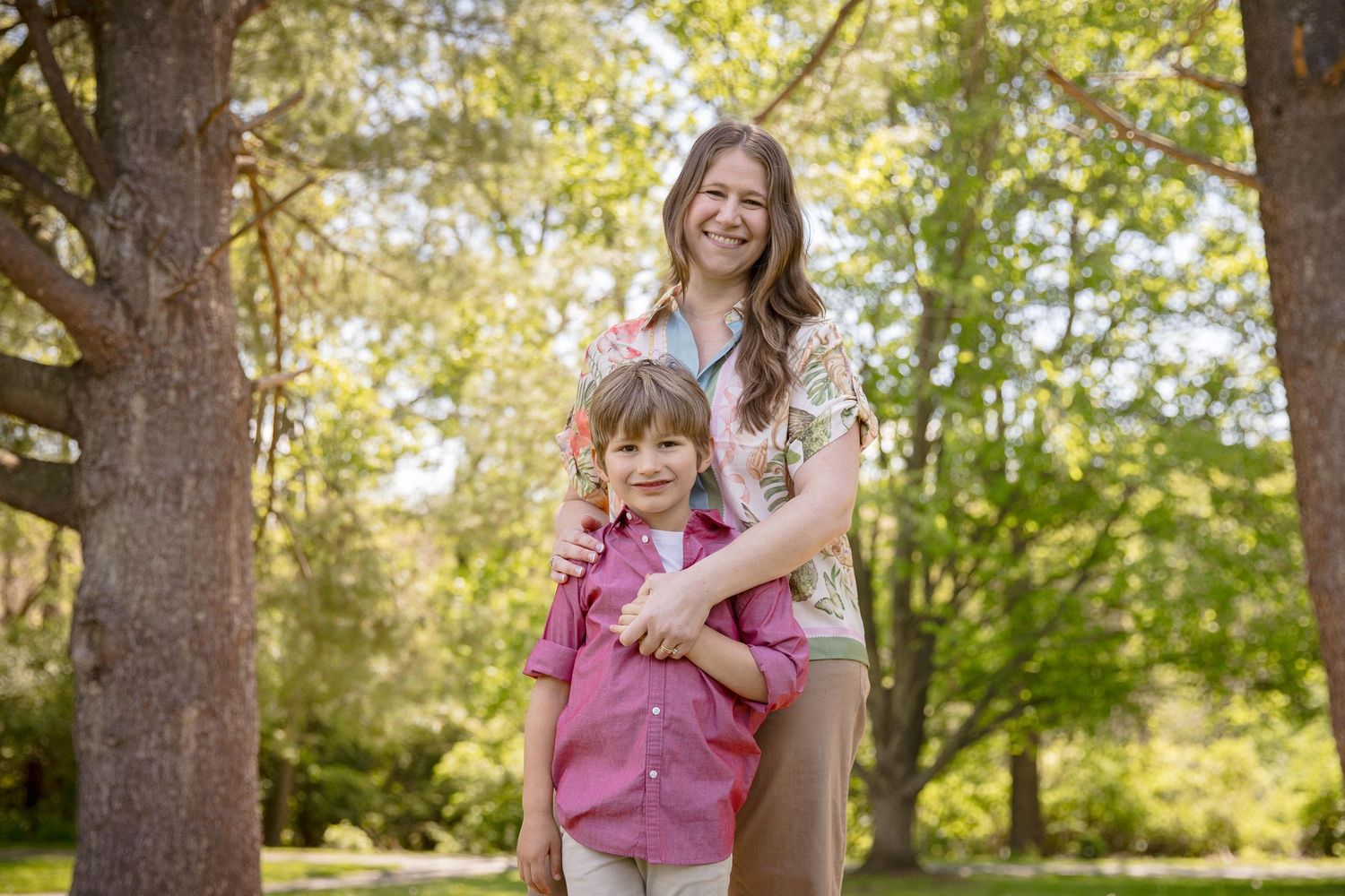 A woman and young boy smile together outdoors among green trees on a sunny day.