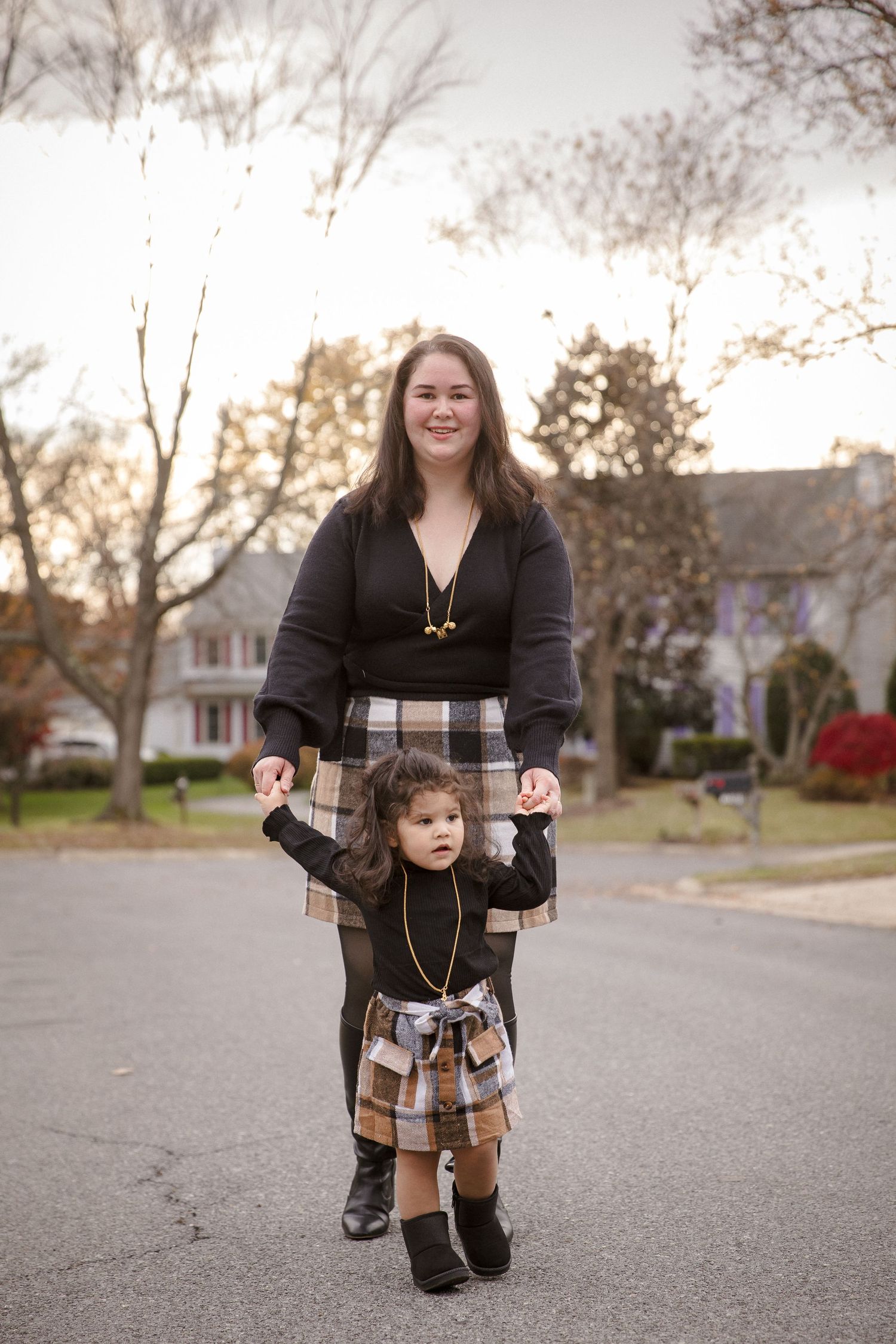 Mother and toddler in matching plaid skirts holding hands on a suburban street during autumn.