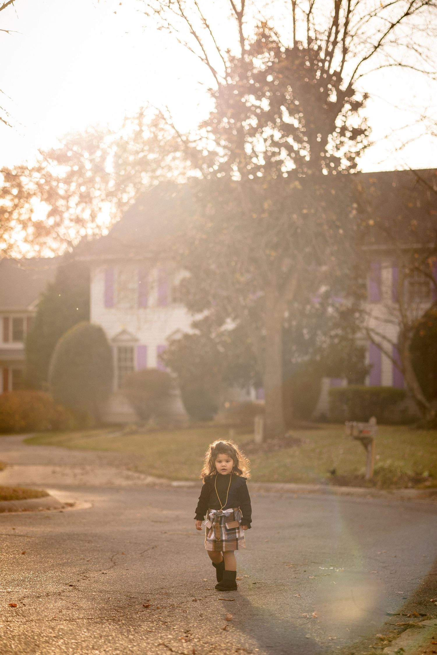 Young girl in plaid skirt walks alone on suburban street bathed in golden autumn sunlight.
