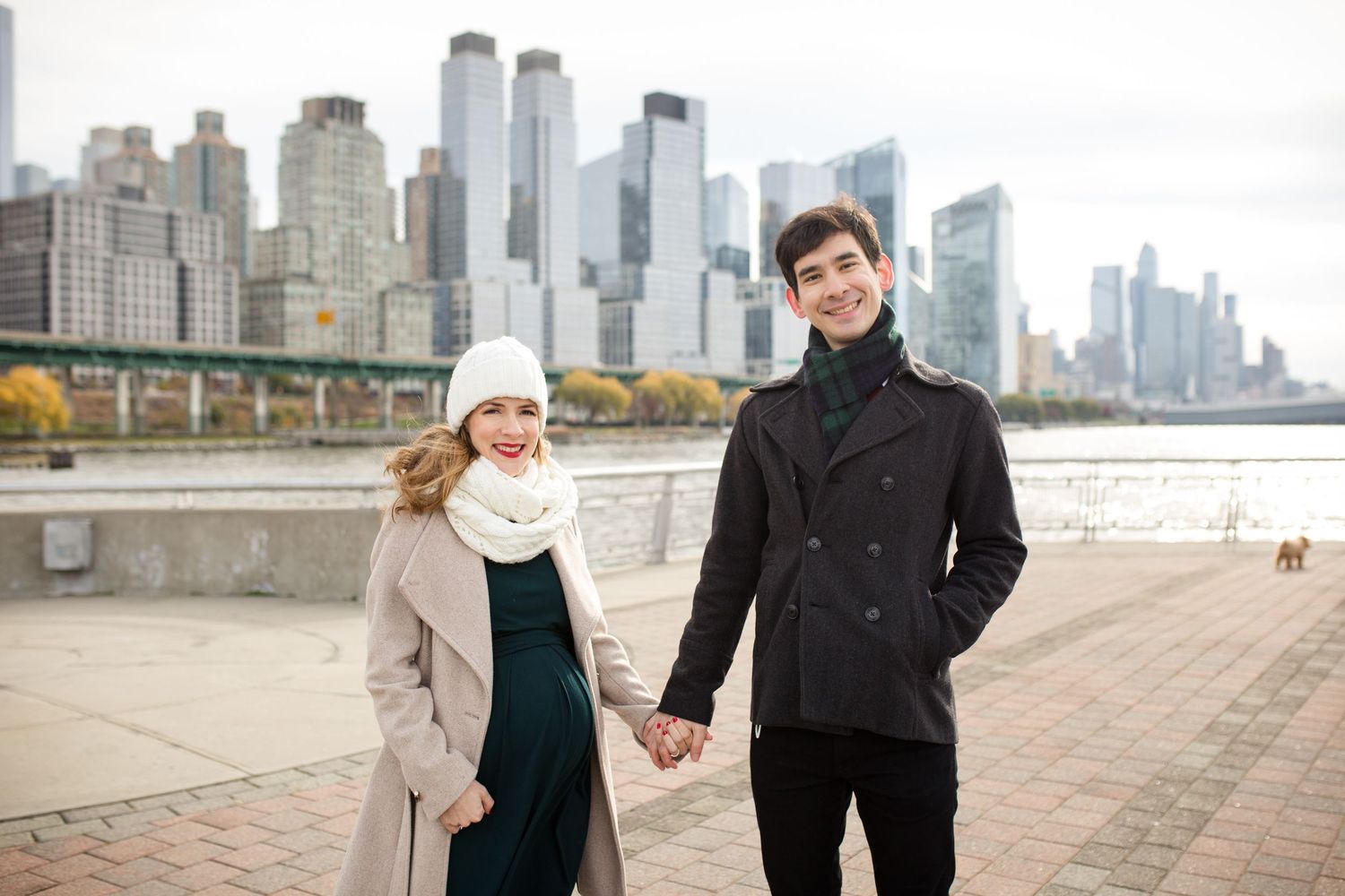 Couple holding hands by NYC waterfront skyline, woman in beige coat and white hat, man in black peacoat.