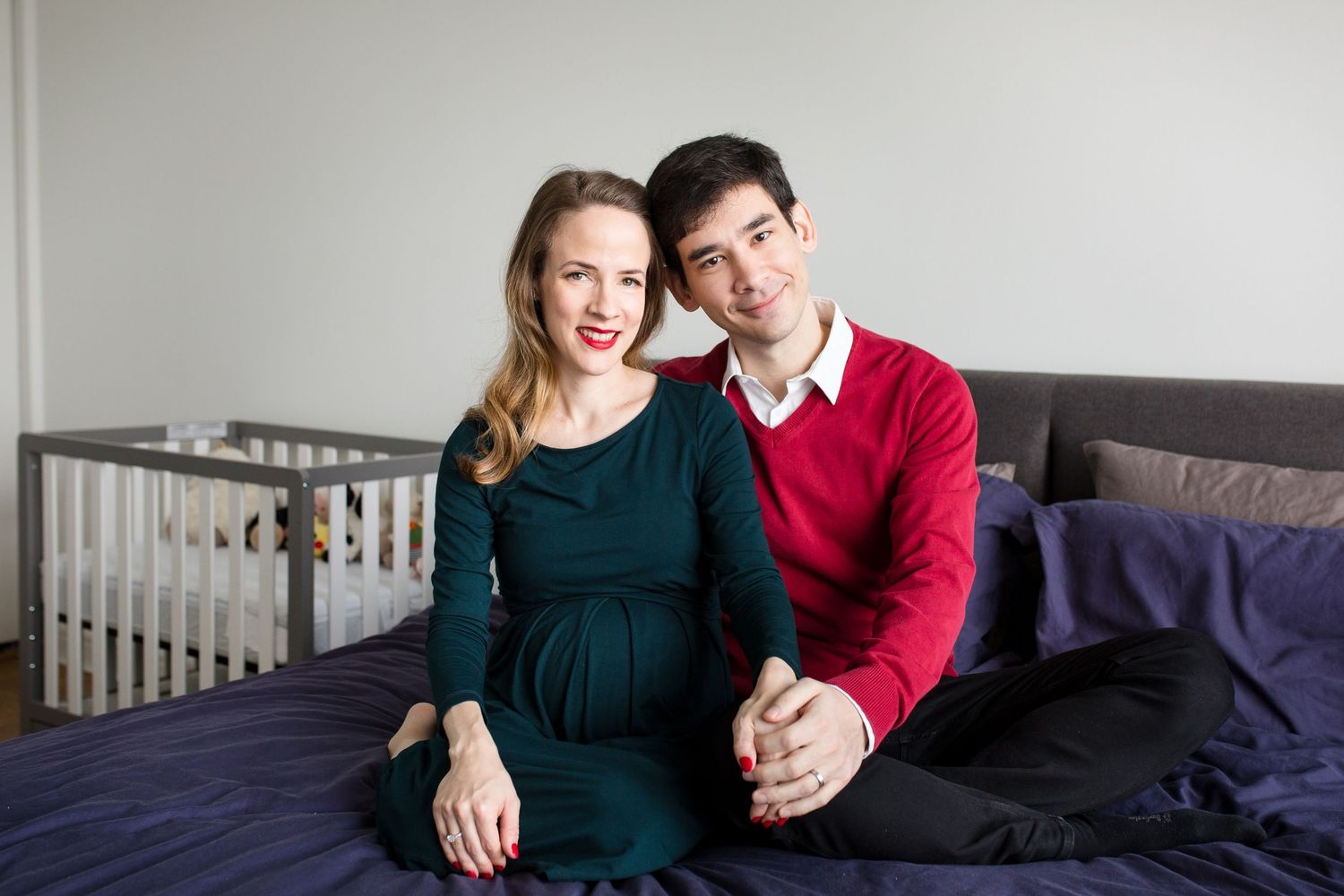 Pregnant woman in green dress and man in red sweater sitting together on bed near baby crib.