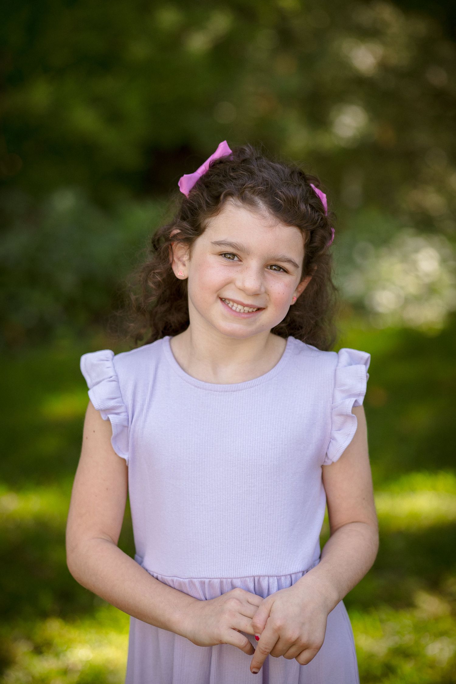 Smiling young girl with curly hair and pink bow wearing a lavender ruffle dress outdoors.
