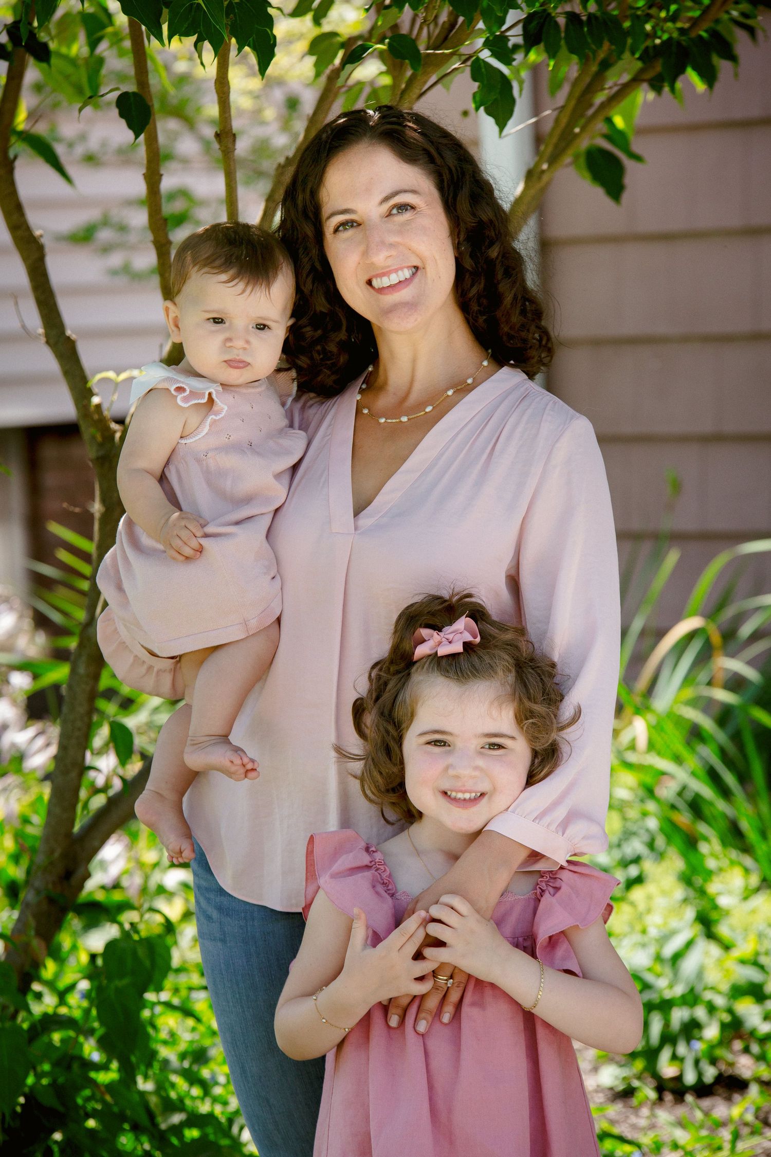 Smiling mother in pink blouse holding baby while older daughter hugs her in a garden.