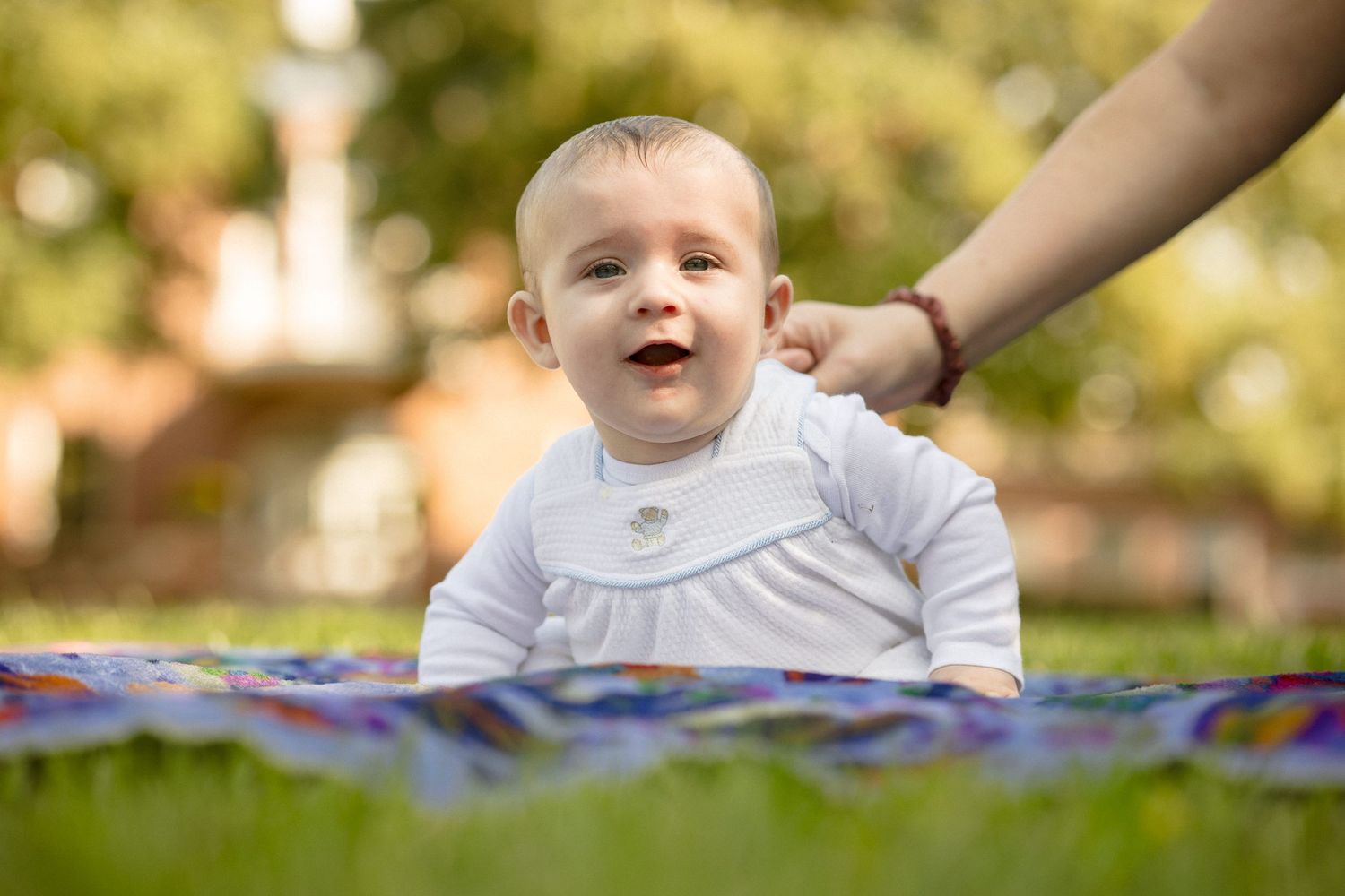 Happy smiling baby in white outfit lying on colorful blanket outdoors in sunny park.