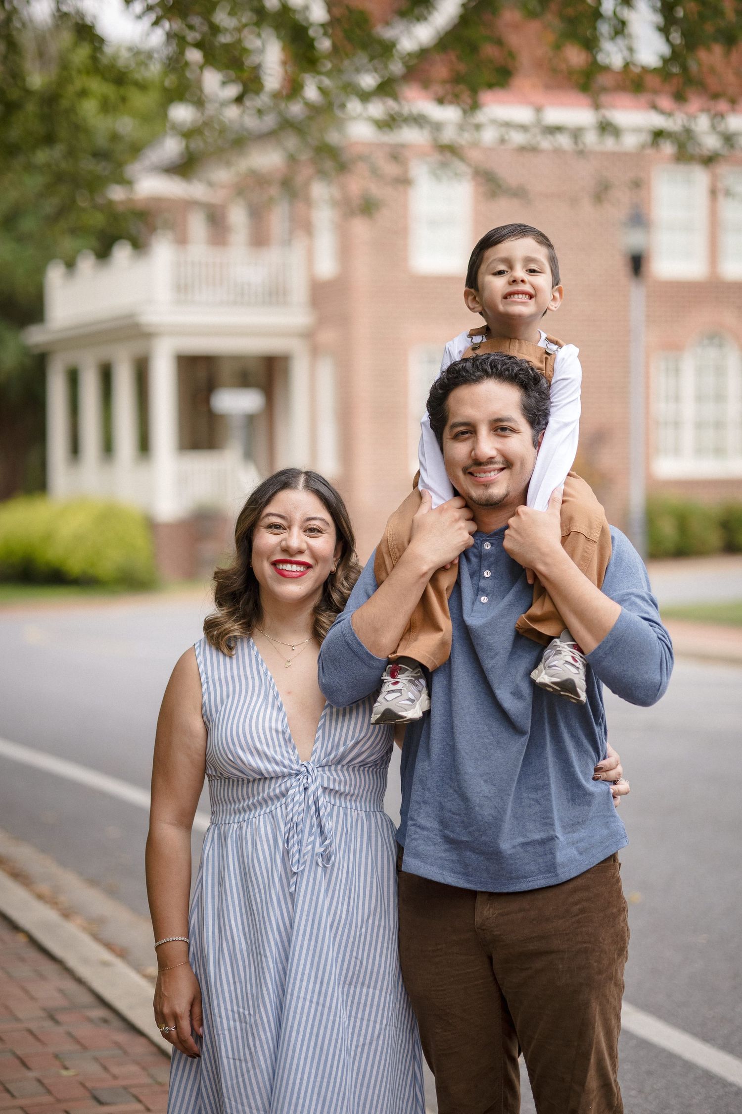A happy family of three poses outdoors, with a young child sitting on dad's shoulders beside a smiling mom in a striped dress.