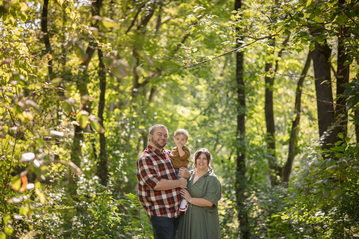 Family of three poses in lush green forest, dad in plaid shirt holds child while mom wears sage green dress.