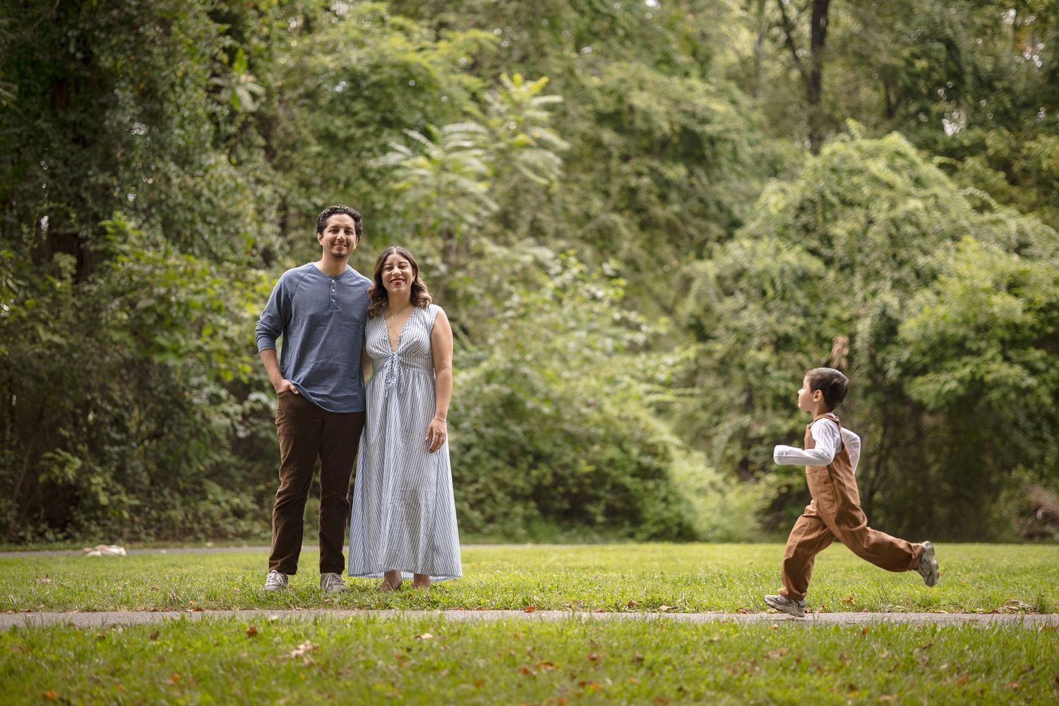 A couple stands smiling in a lush green park while a young child runs toward them on a sunny day.