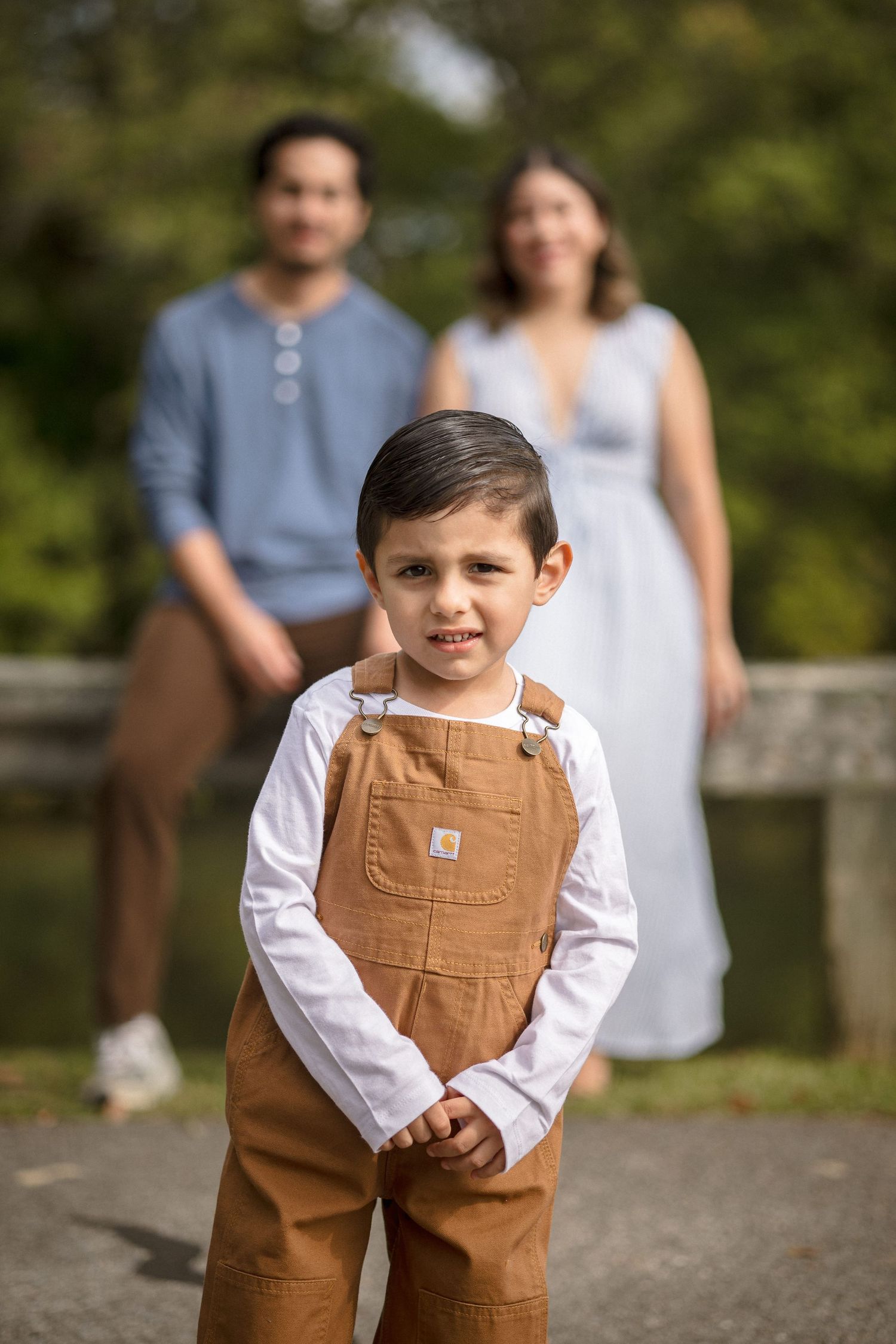 Young boy in Carhartt overalls smiles at camera with parents posing in blurred background outdoors.