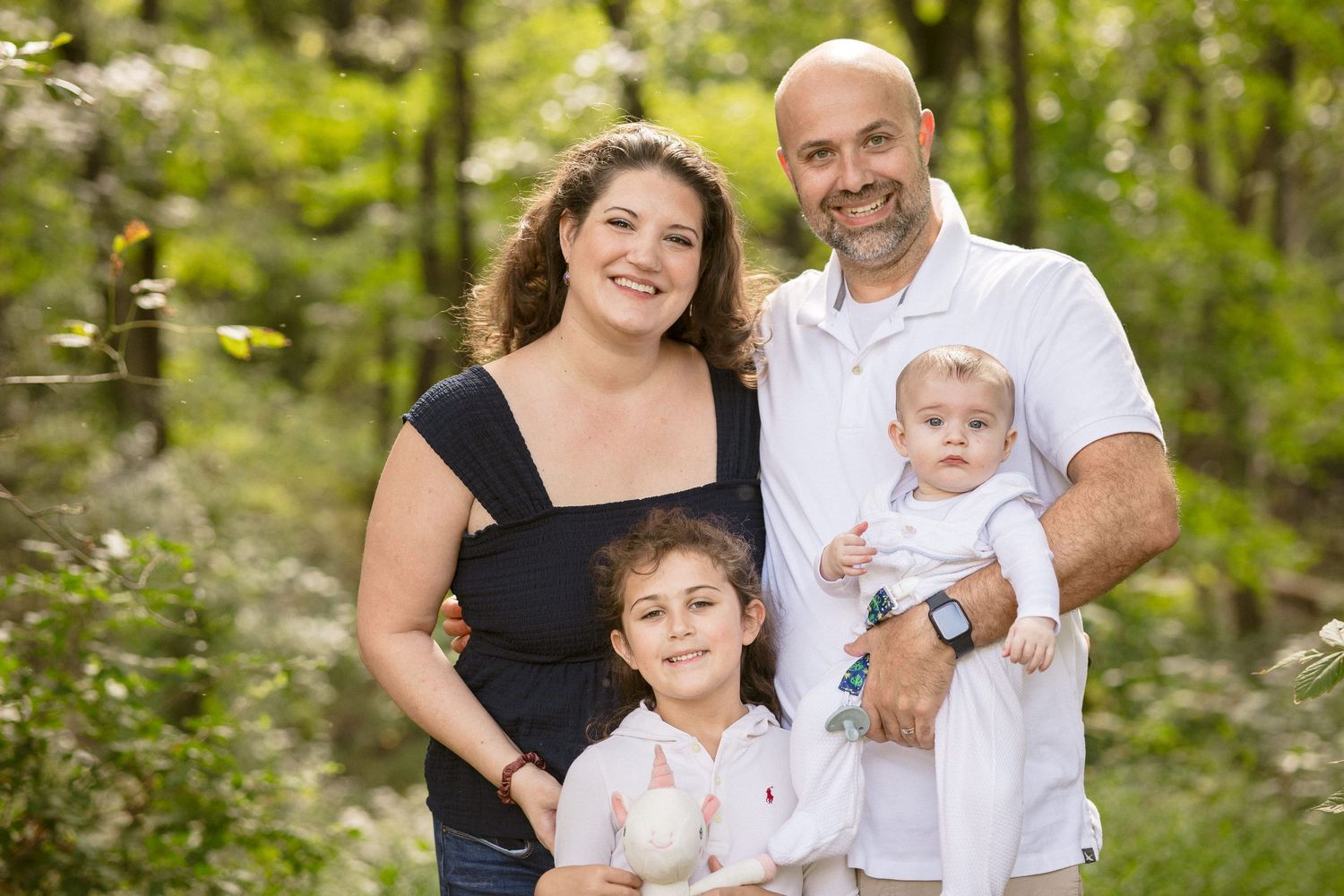 Happy family of four posing outdoors in lush green forest setting, parents smiling with young daughter and baby.