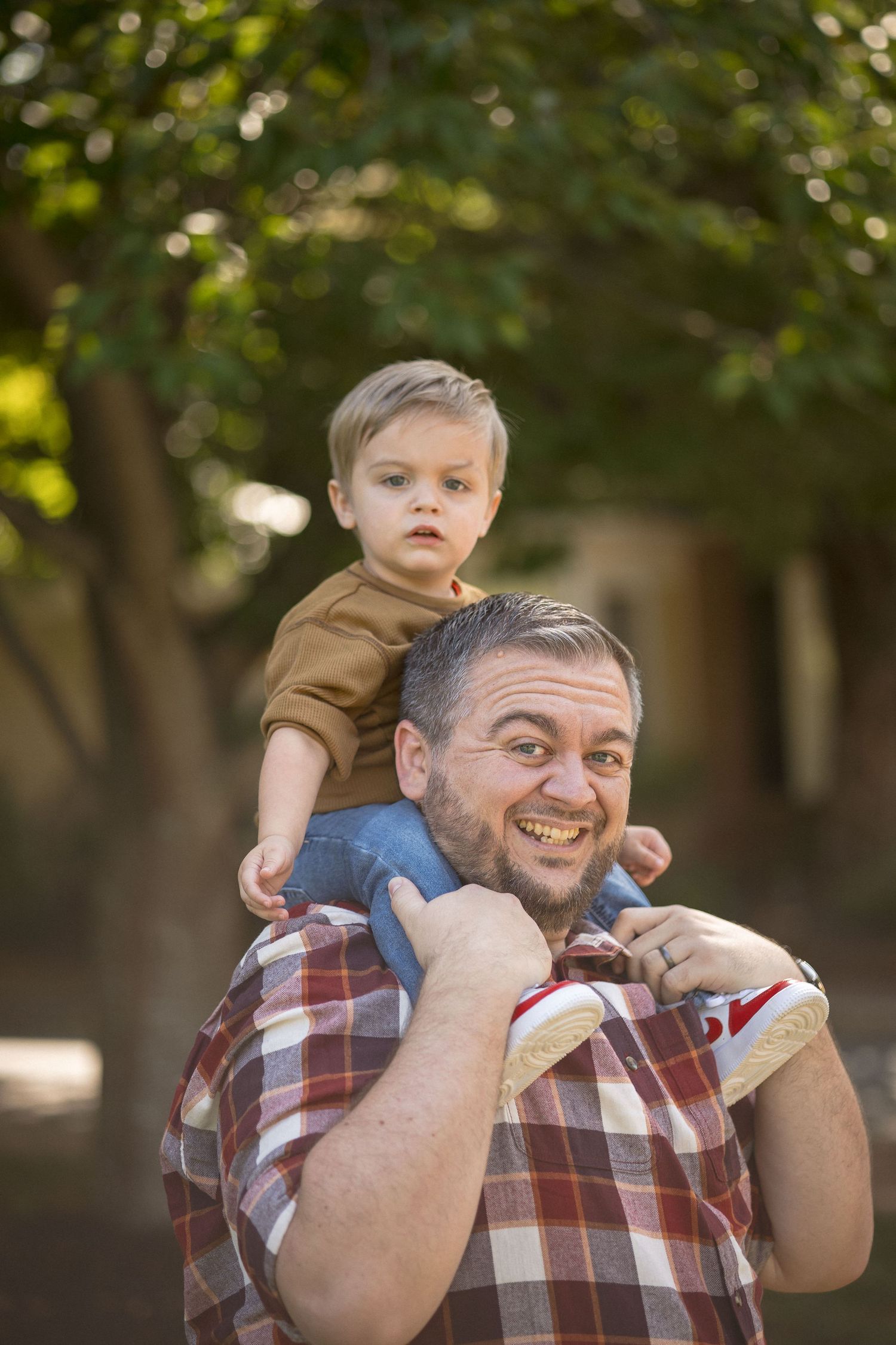 A smiling father carries his young toddler son on his shoulders outdoors in a park with green trees.