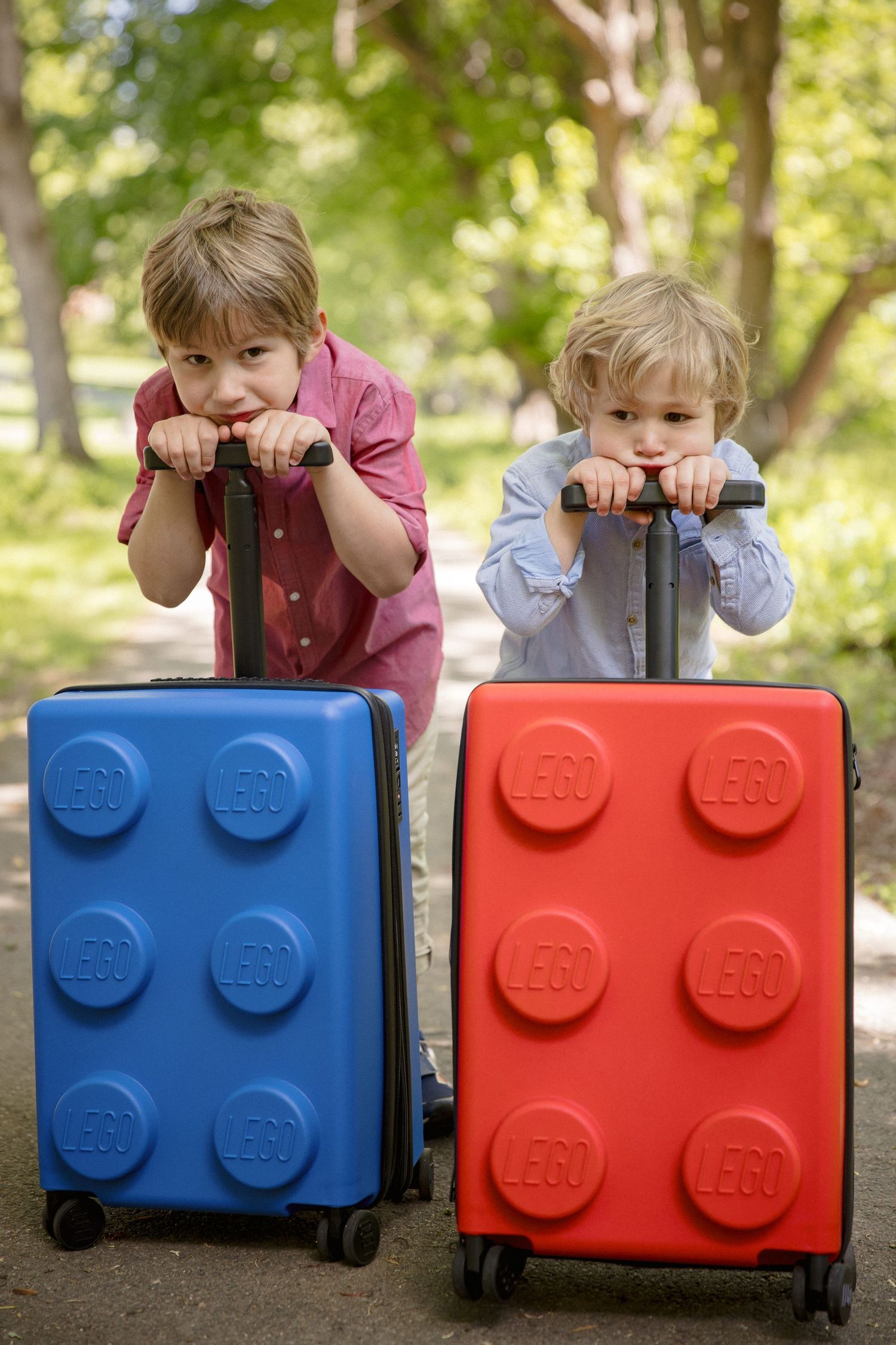 Two young boys leaning on colorful LEGO-themed blue and red suitcases outdoors in a sunny park.
