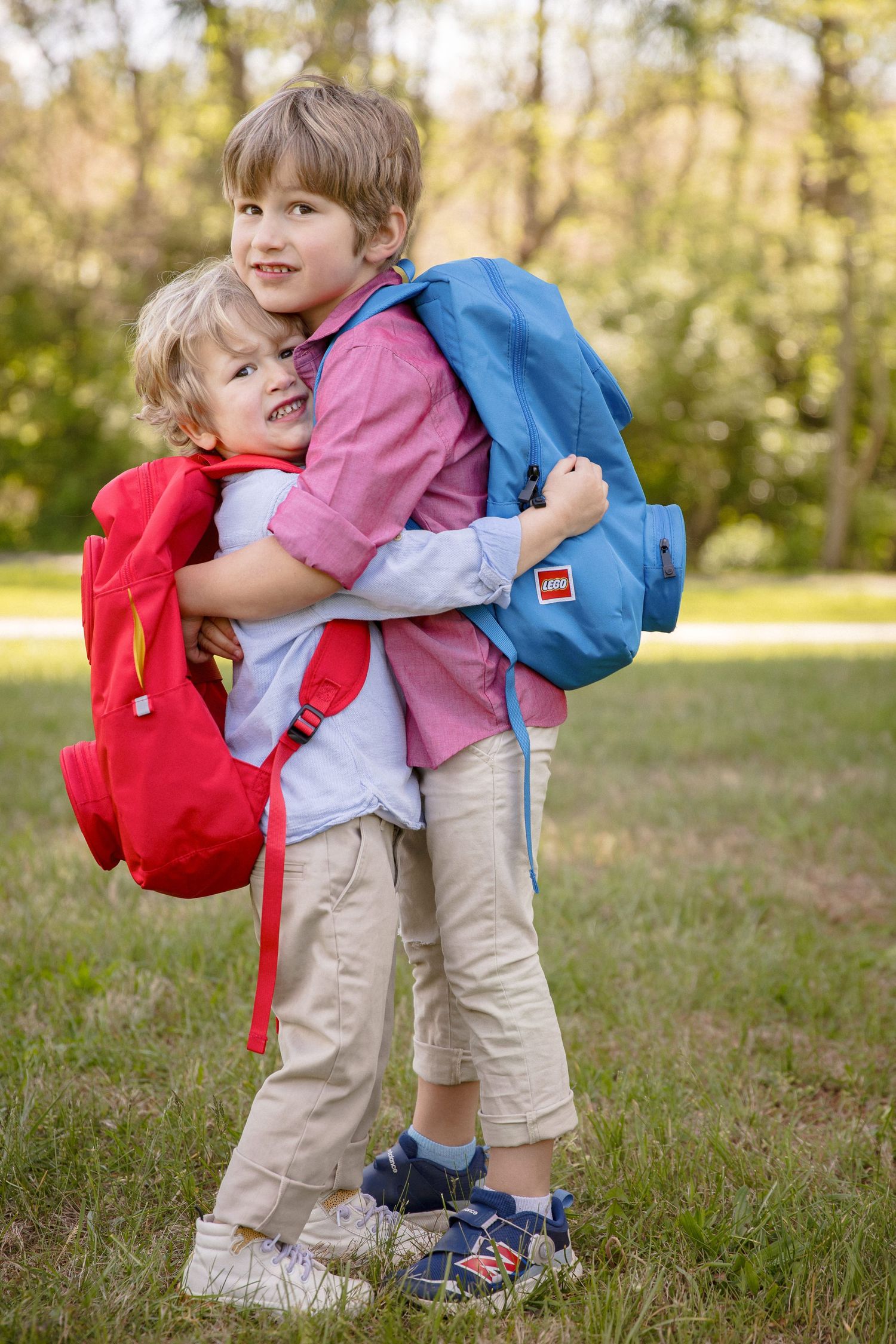 Two young boys with backpacks hugging outdoors, smiling happily in a park setting on a sunny day.