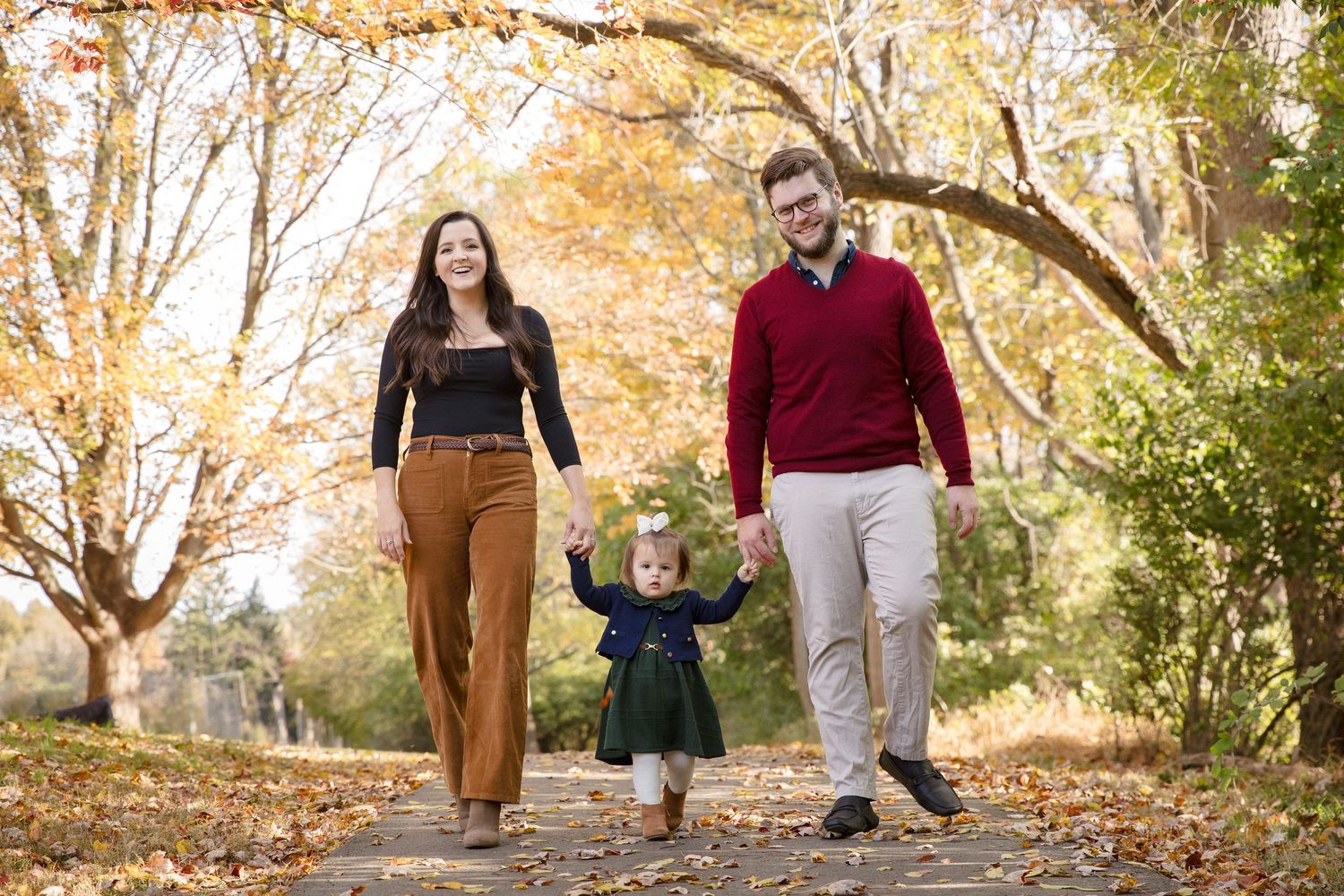 Young family of three holding hands walking down a tree-lined path surrounded by golden autumn leaves.