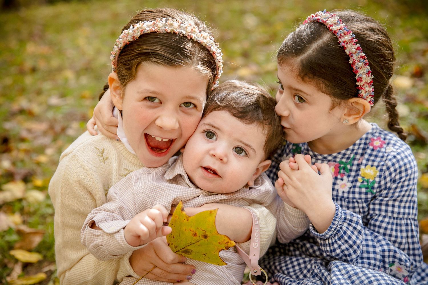 Two young girls hug a smiling baby holding a yellow leaf in an autumn park setting.