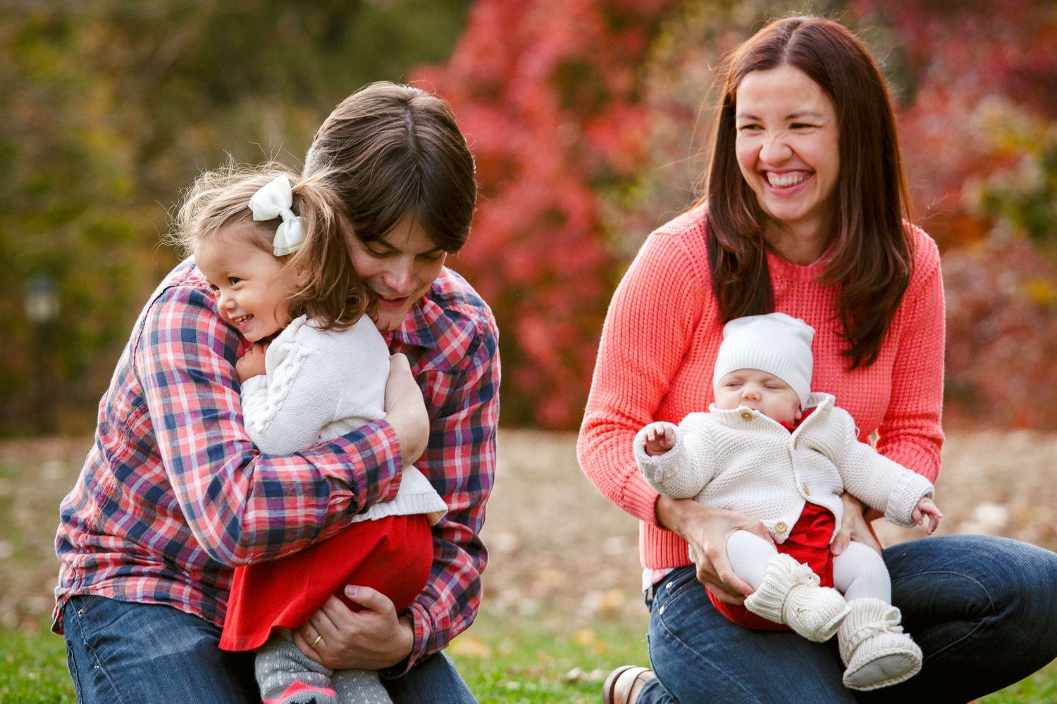 Two smiling mothers outdoors in autumn, one hugging a toddler girl and the other holding a newborn baby.
