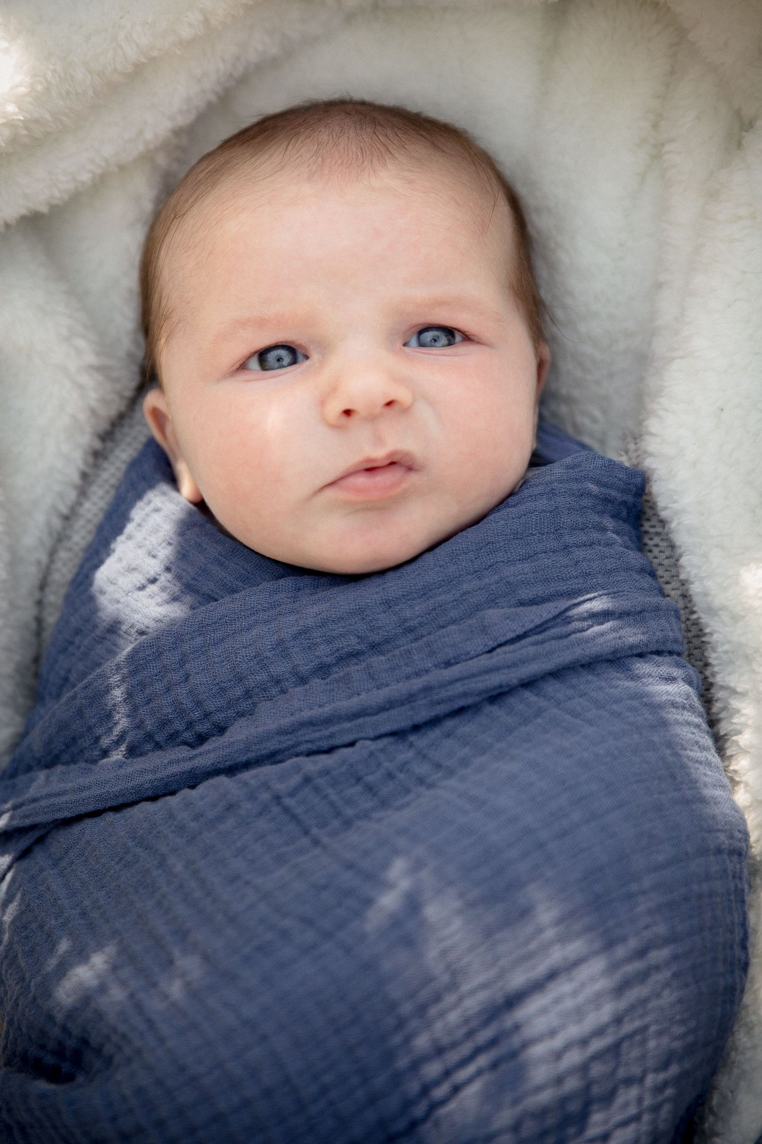 Newborn baby swaddled in blue muslin blanket on white fleece.