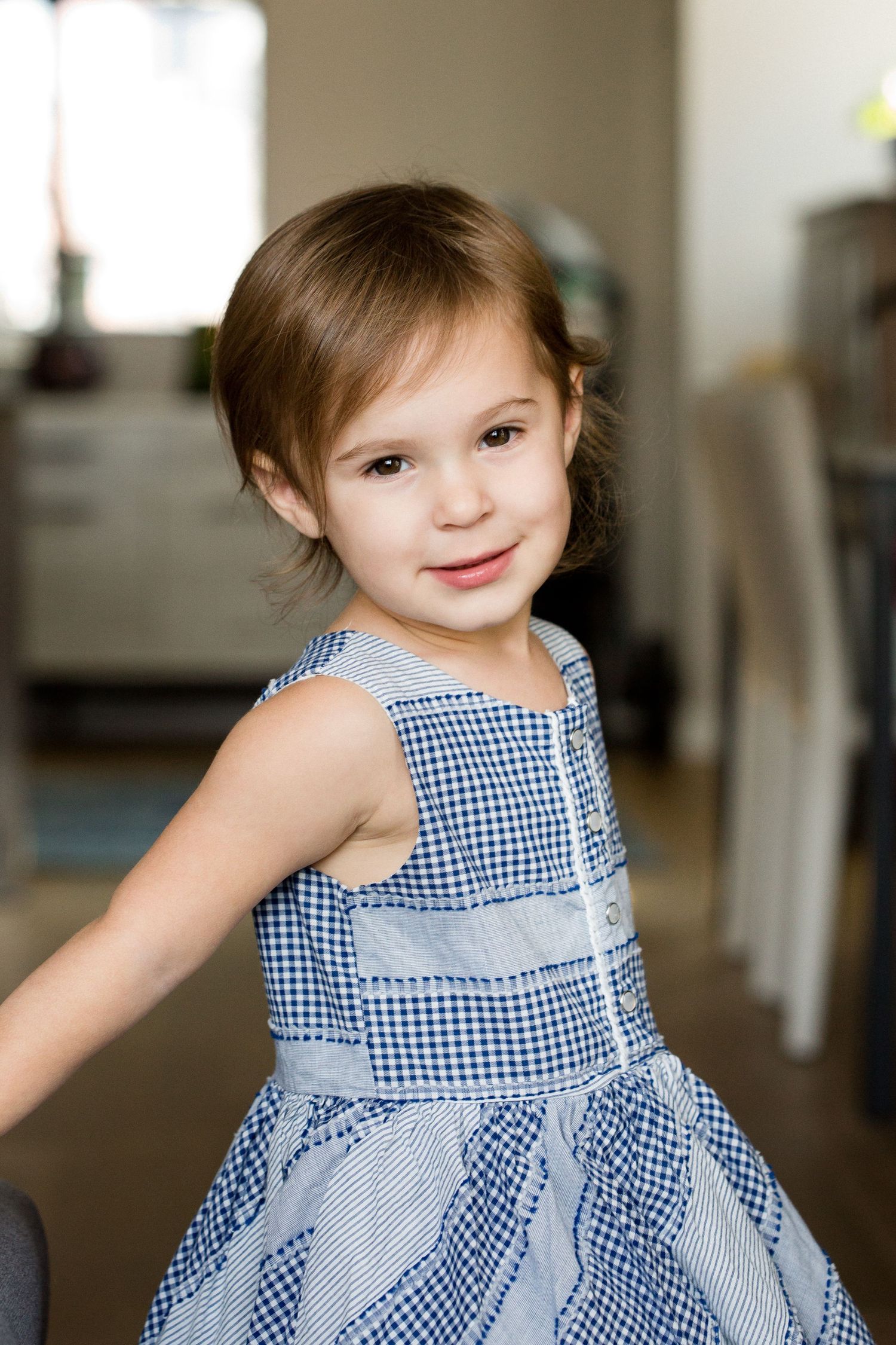 Cute toddler girl in blue gingham dress smiling over shoulder in bright indoor home setting.