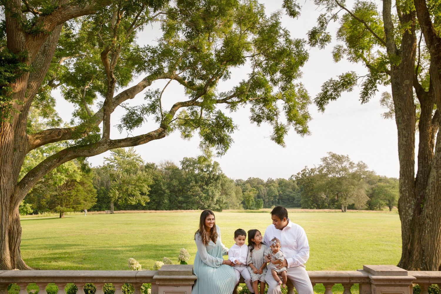 Family of four enjoying a sunny day together on a stone terrace overlooking a lush green park.