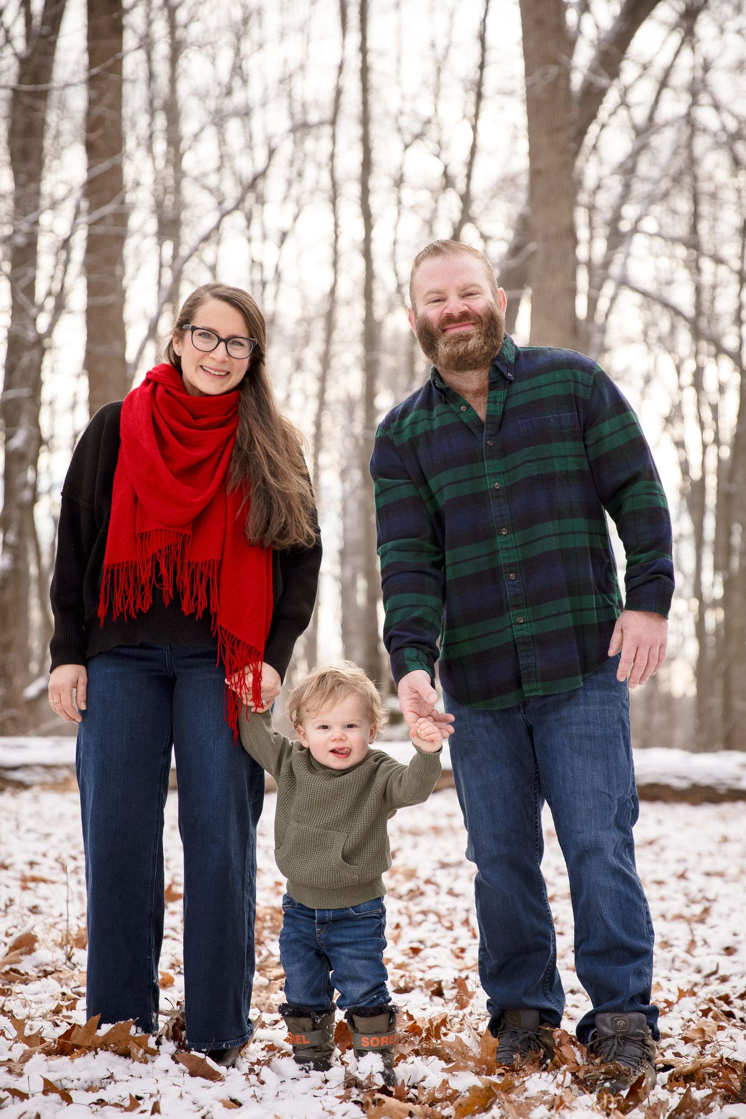 Smiling family of three holds hands in a snowy winter forest with fallen leaves.