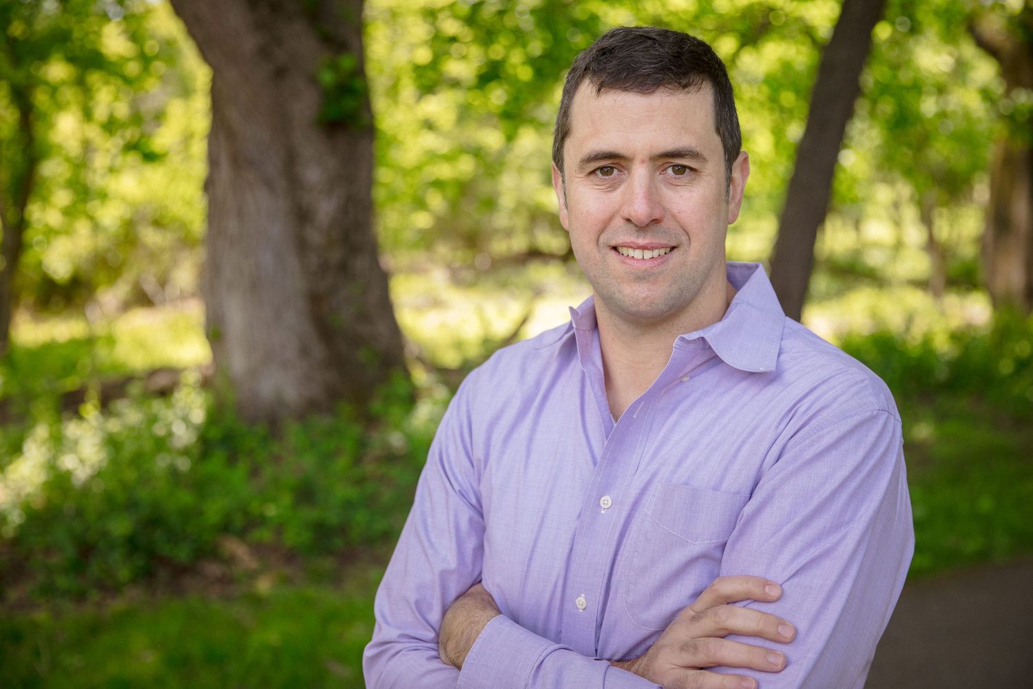 Man in purple shirt smiling with arms crossed outdoors among green trees in a professional portrait photo.