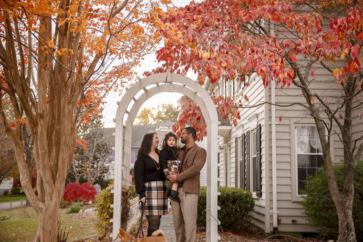 Family of three poses under white garden arbor surrounded by vibrant red autumn foliage outside a home.