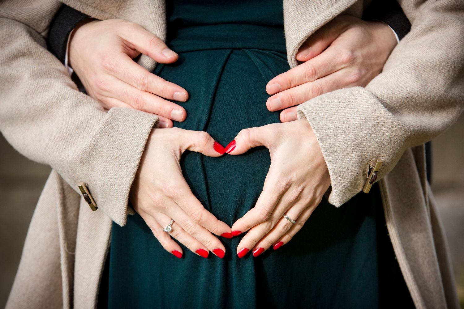 Couple forming heart shape with hands on pregnant belly, woman with red nails wearing engagement ring and teal dress.