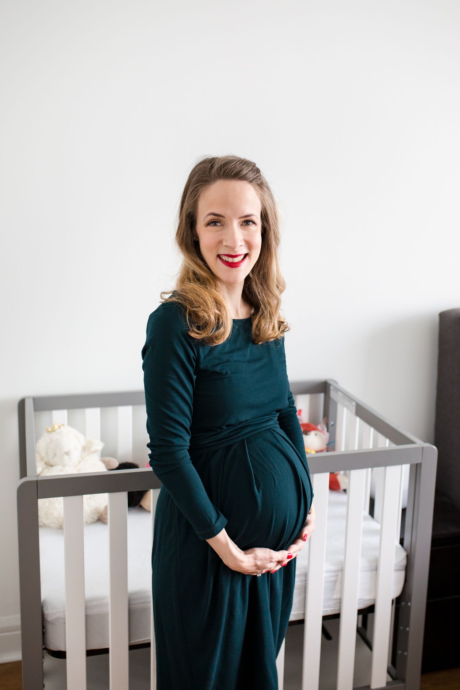 Smiling pregnant woman in dark green dress cradling baby bump, standing in front of a gray nursery crib.