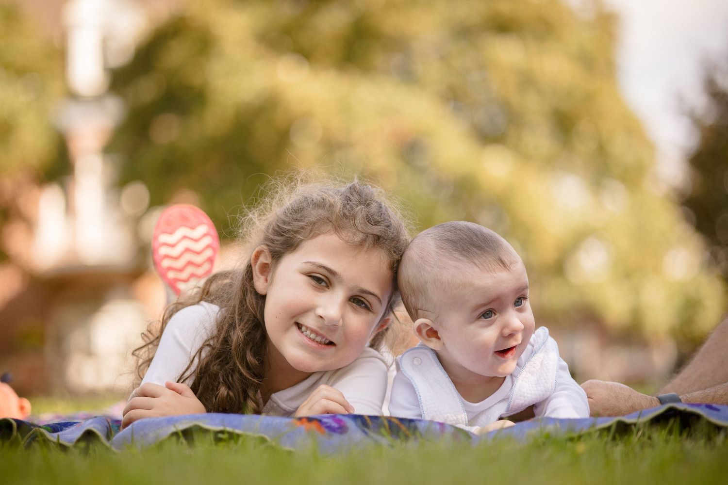 A smiling girl and baby lying on grass in a sunny park, enjoying a peaceful outdoor moment together.