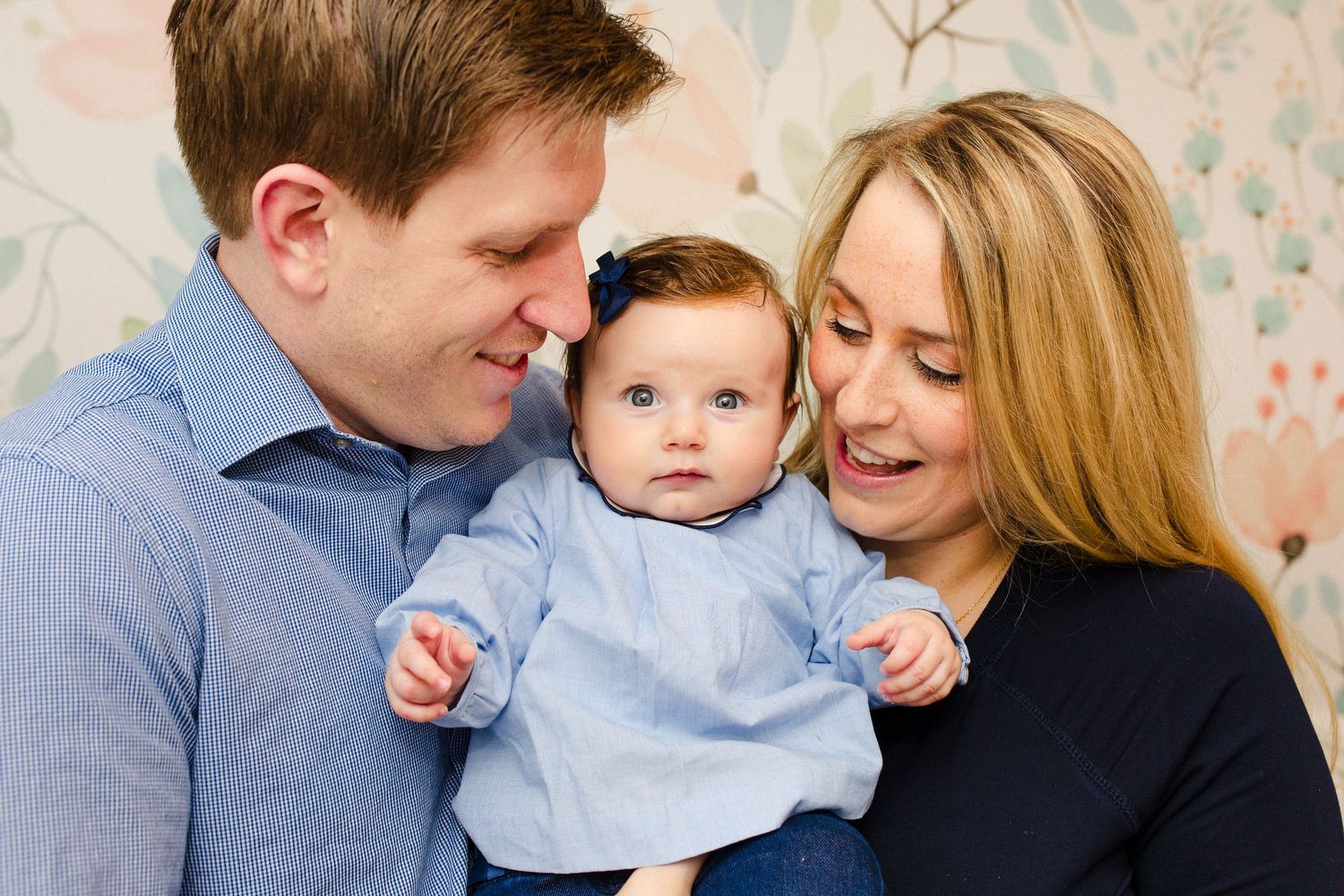 Happy young parents holding their baby girl in blue outfit against floral wallpaper background.