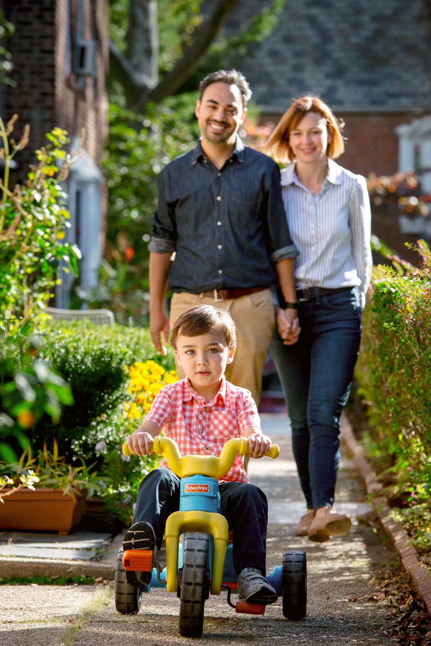 Young boy riding tricycle with parents walking behind in garden.