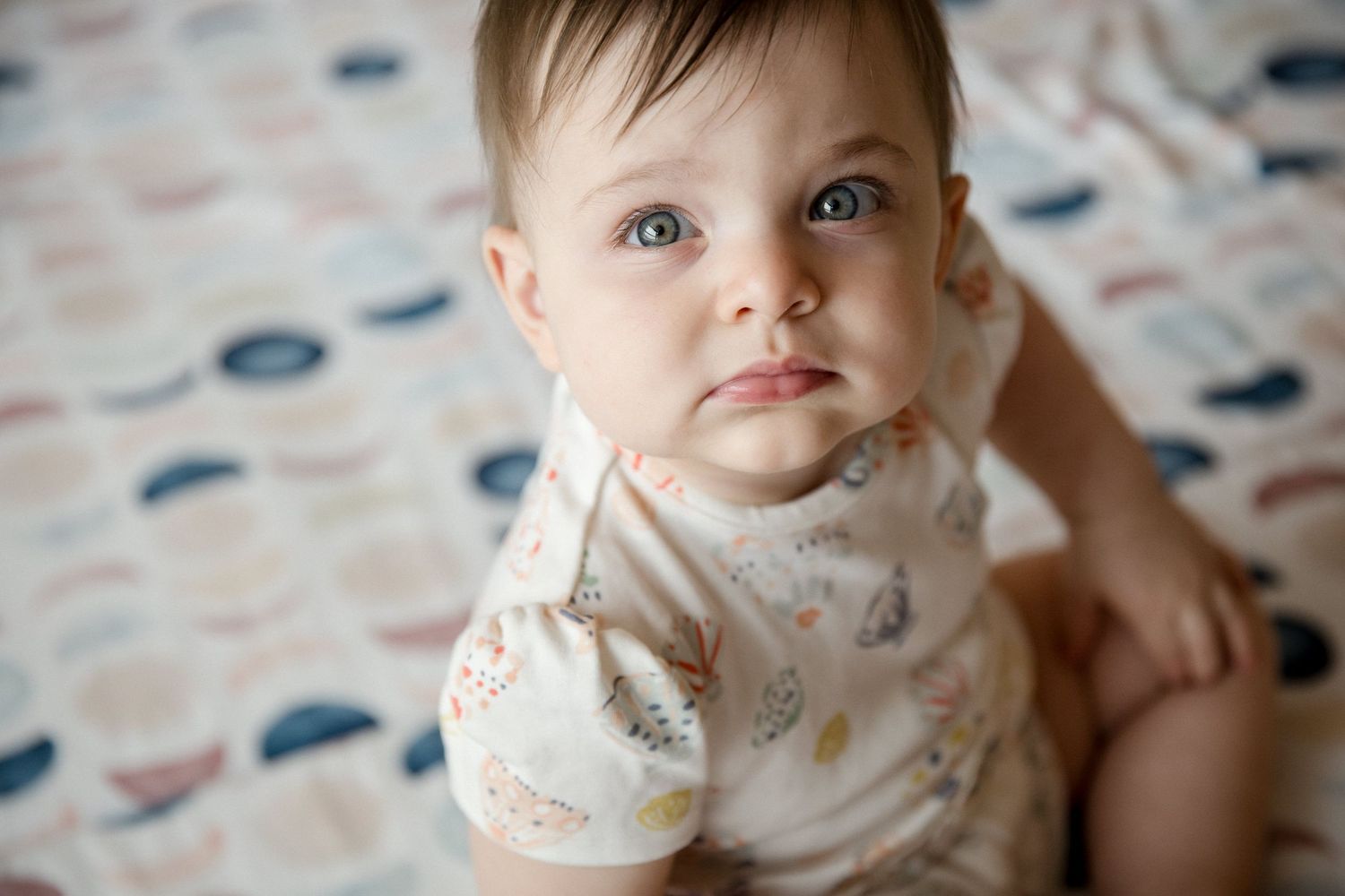 Adorable baby with blue eyes wearing a floral onesie looking up curiously while sitting on a patterned blanket.