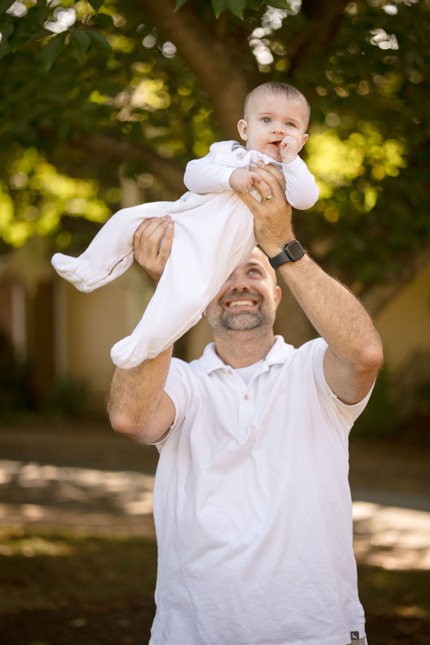 A smiling man in white shirt lifts a happy baby overhead outdoors, surrounded by green trees.
