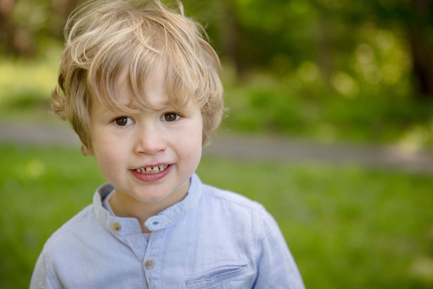 Smiling blond toddler boy in a light blue shirt outdoors in a green park setting.