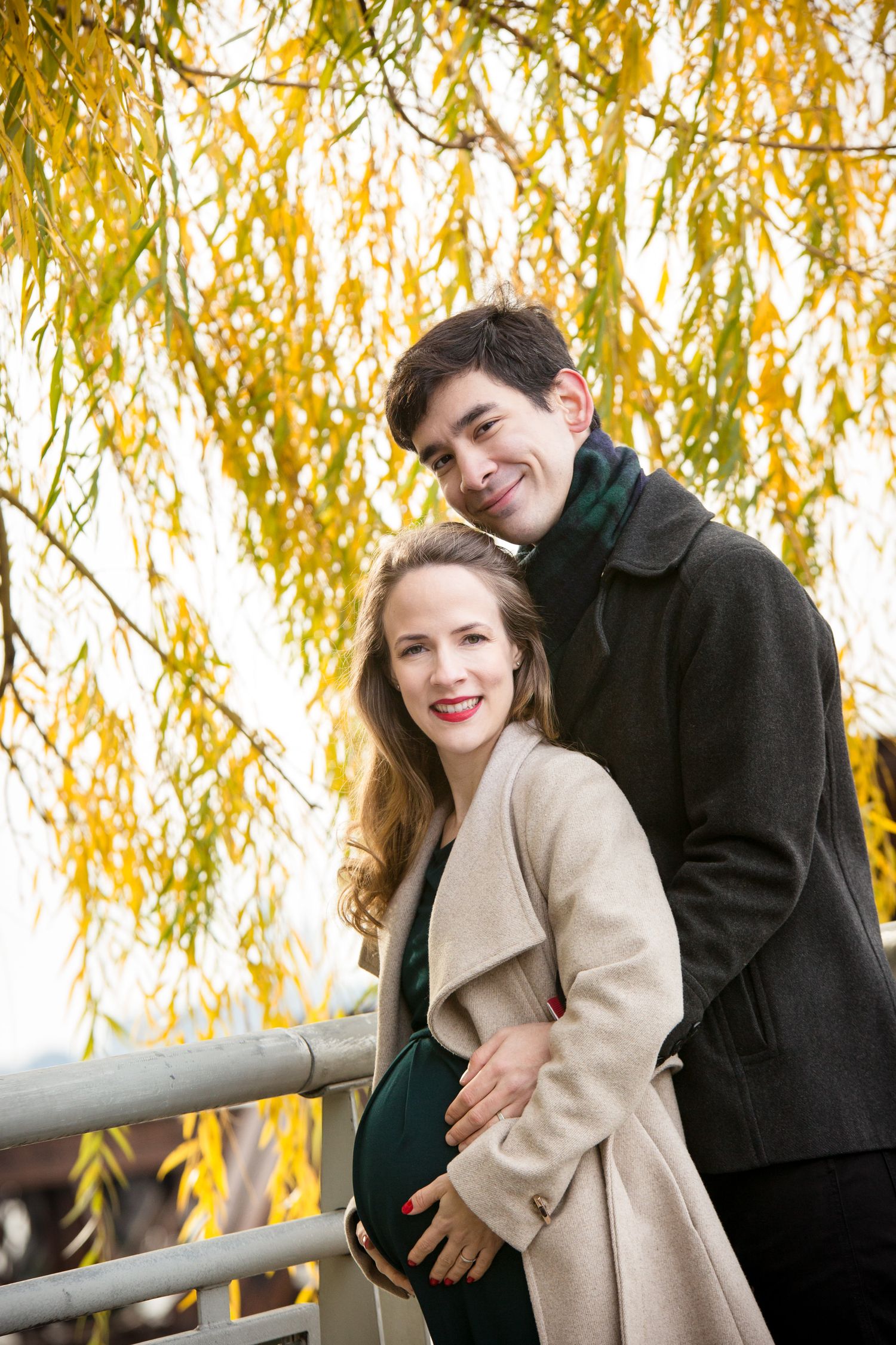 Happy couple posing outdoors in autumn, pregnant woman in beige coat with partner embracing her baby bump near golden willow tree.