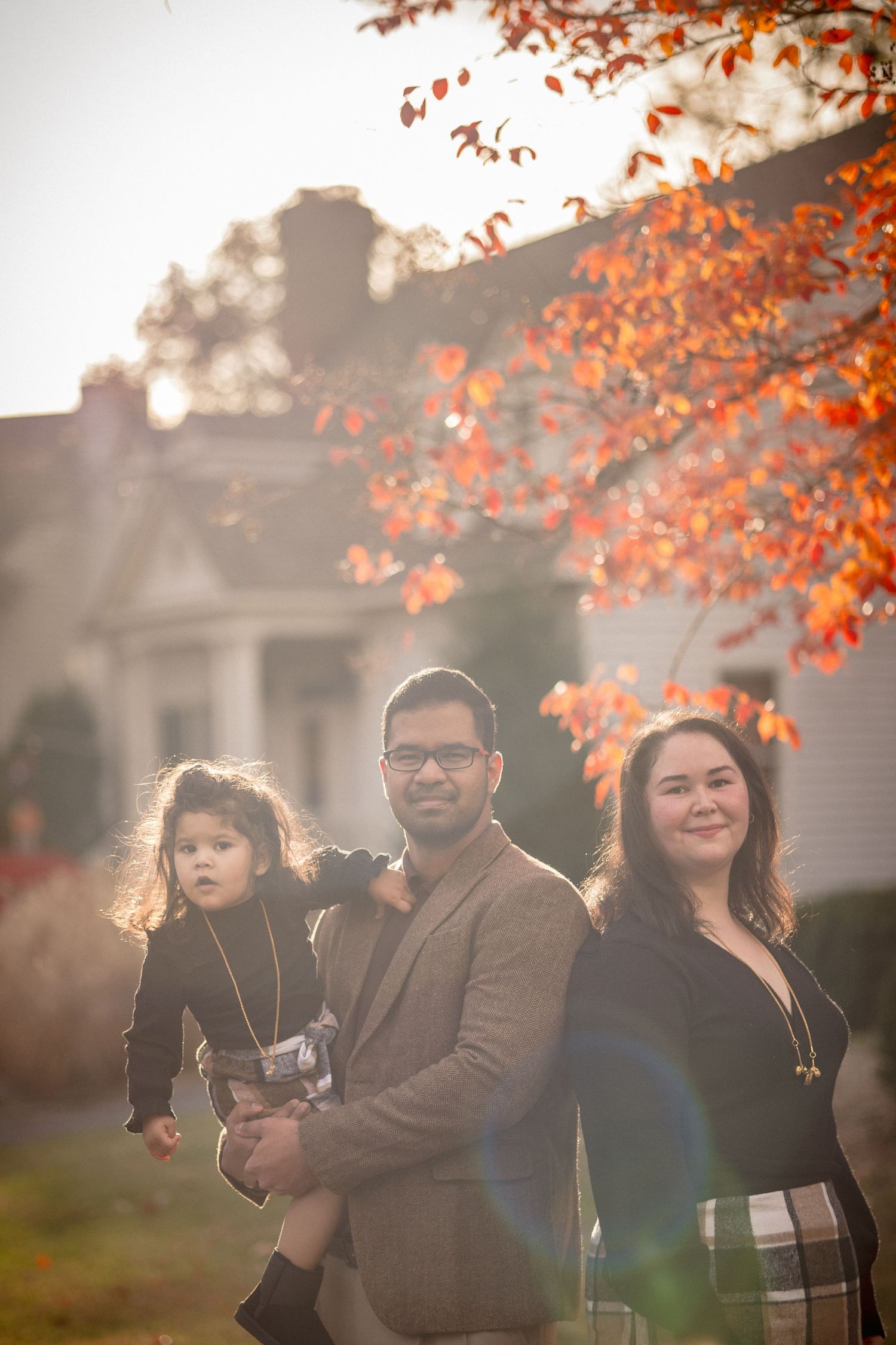 Family of three posing outdoors in autumn, father holding toddler beside mother, vibrant orange leaves and warm golden light.